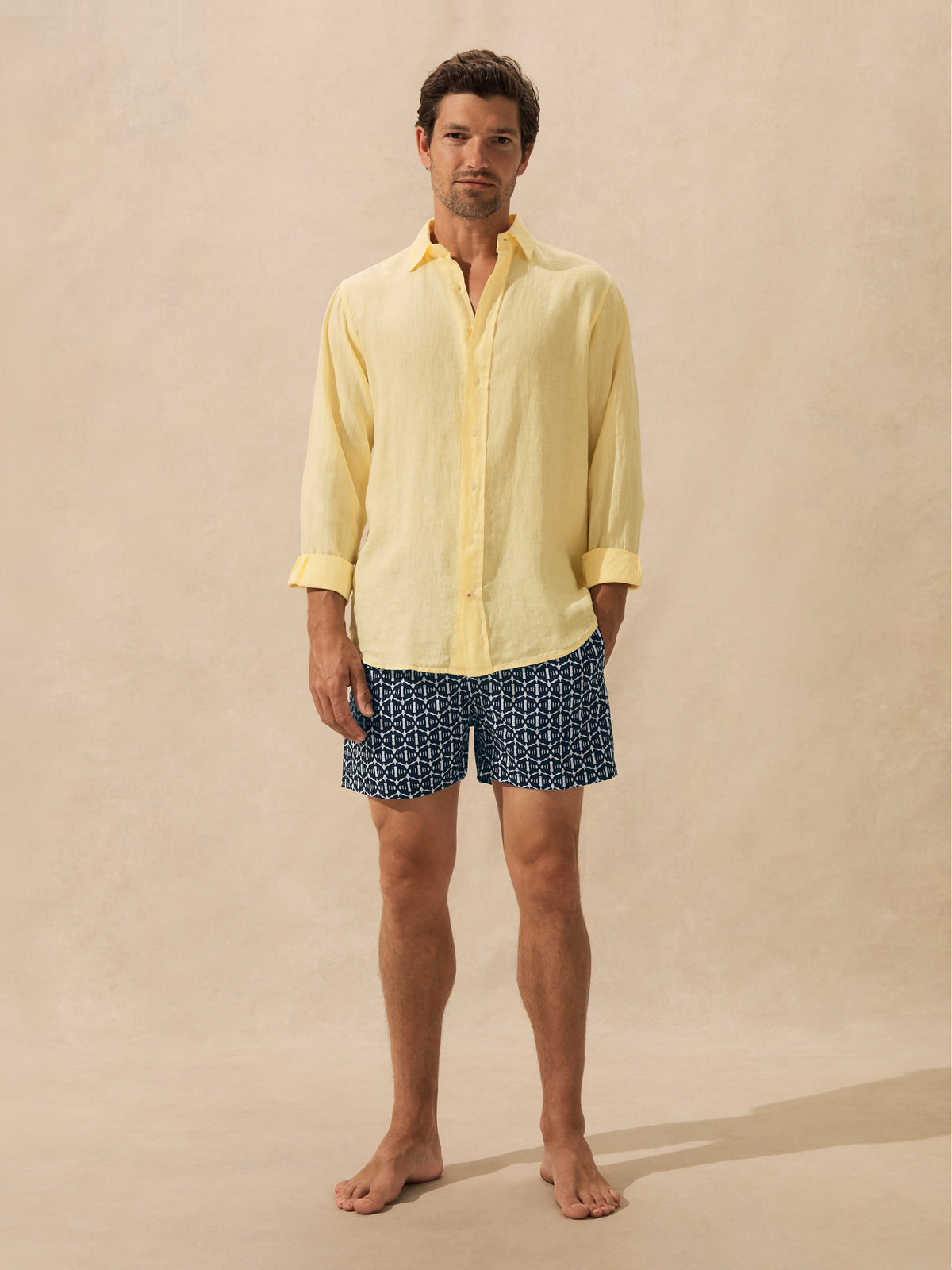 A man stands barefoot against a warm beige studio backdrop, wearing a pale yellow long-sleeve button-up shirt and dark blue patterned shorts.