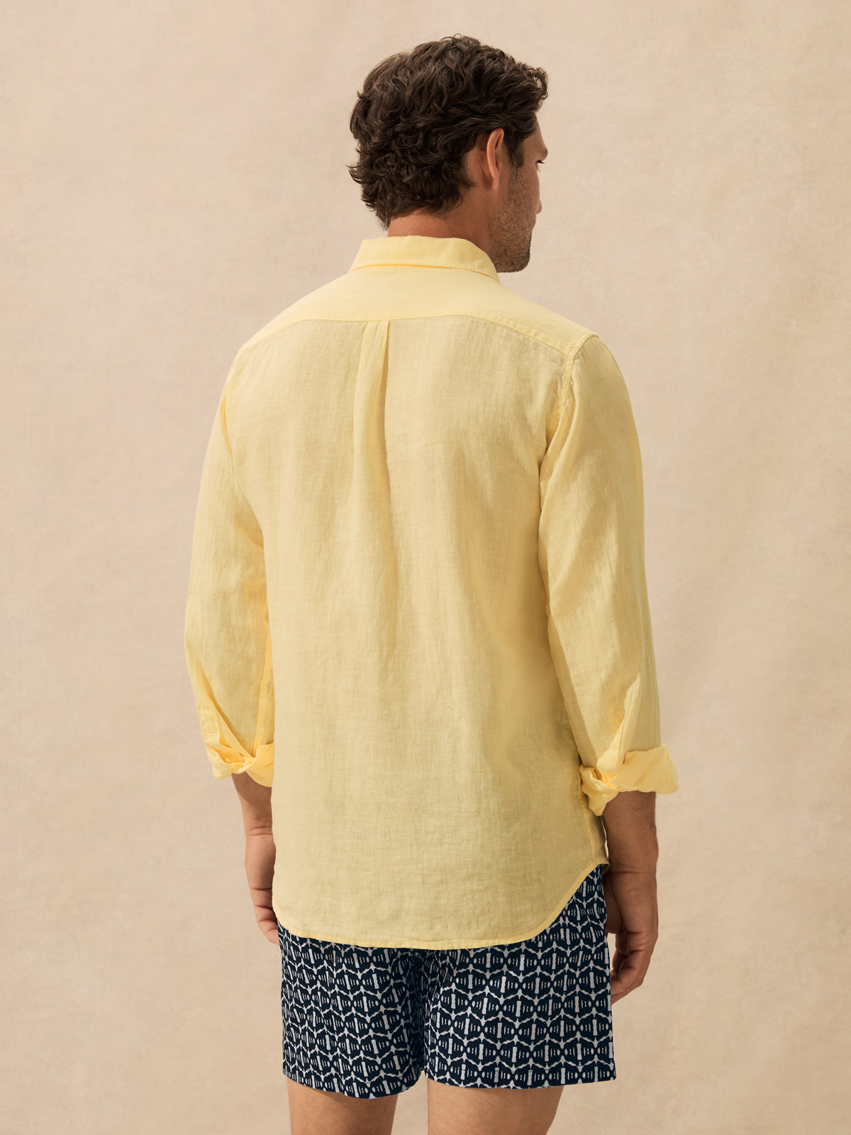 A man viewed from behind wearing a pale yellow linen button-up shirt and navy shorts with a white geometric pattern, standing against a textured beige background.