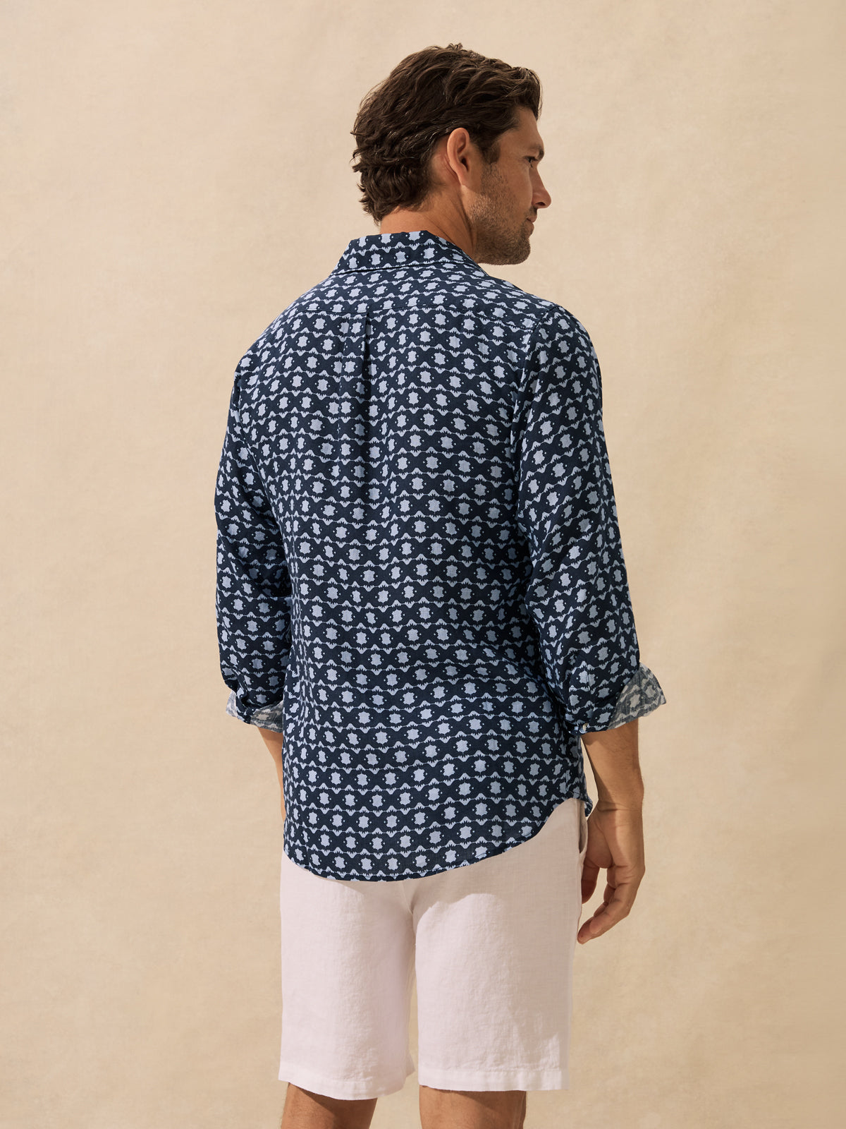 A man shown from the back wearing a navy blue long-sleeve button-up shirt with a white geometric block print pattern, paired with white linen shorts, against a textured beige background.