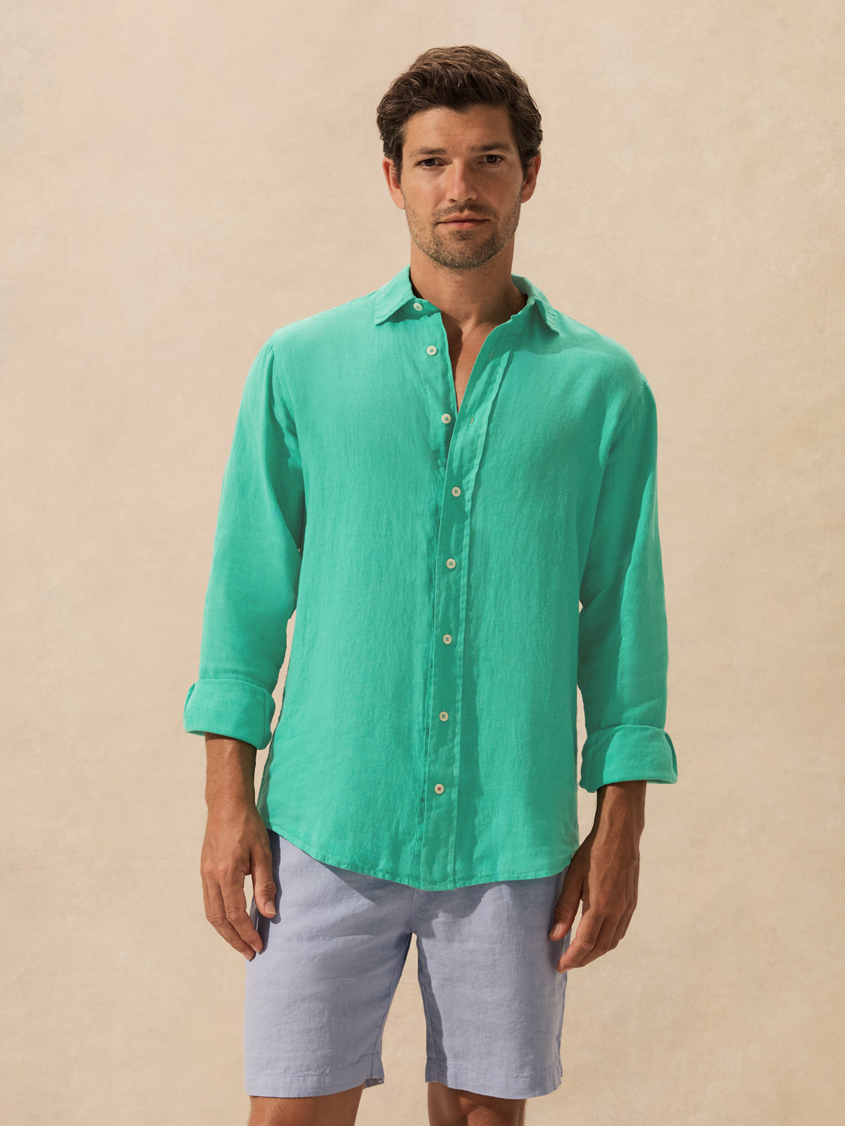 A man wearing a mint green long-sleeve linen button-down shirt and light blue shorts, standing against a neutral beige background.