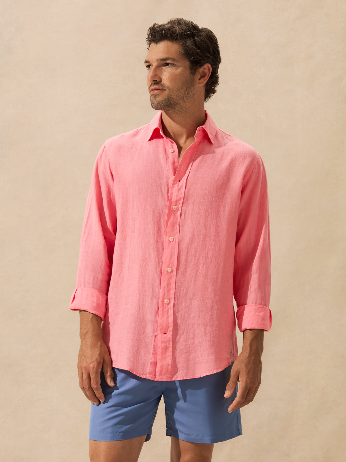 A man wearing a pink long-sleeve linen button-down shirt with rolled-up sleeves standing against a textured background.