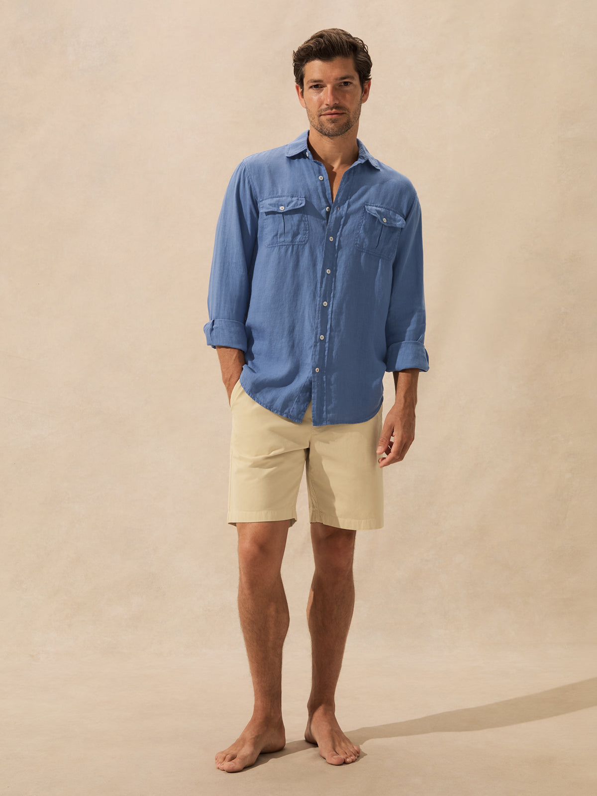 A man stands barefoot against a warm beige studio backdrop, wearing a blue long-sleeve button-up shirt with chest pockets and beige shorts.