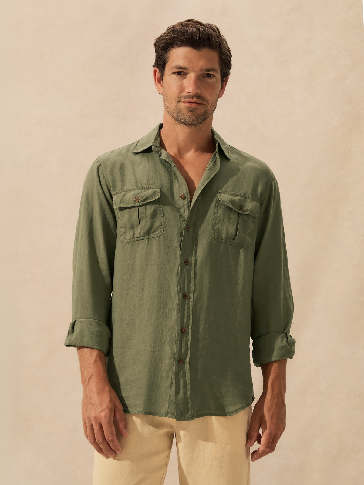 A man wearing an olive green long-sleeve button-up shirt with two chest pockets standing against a textured beige background.