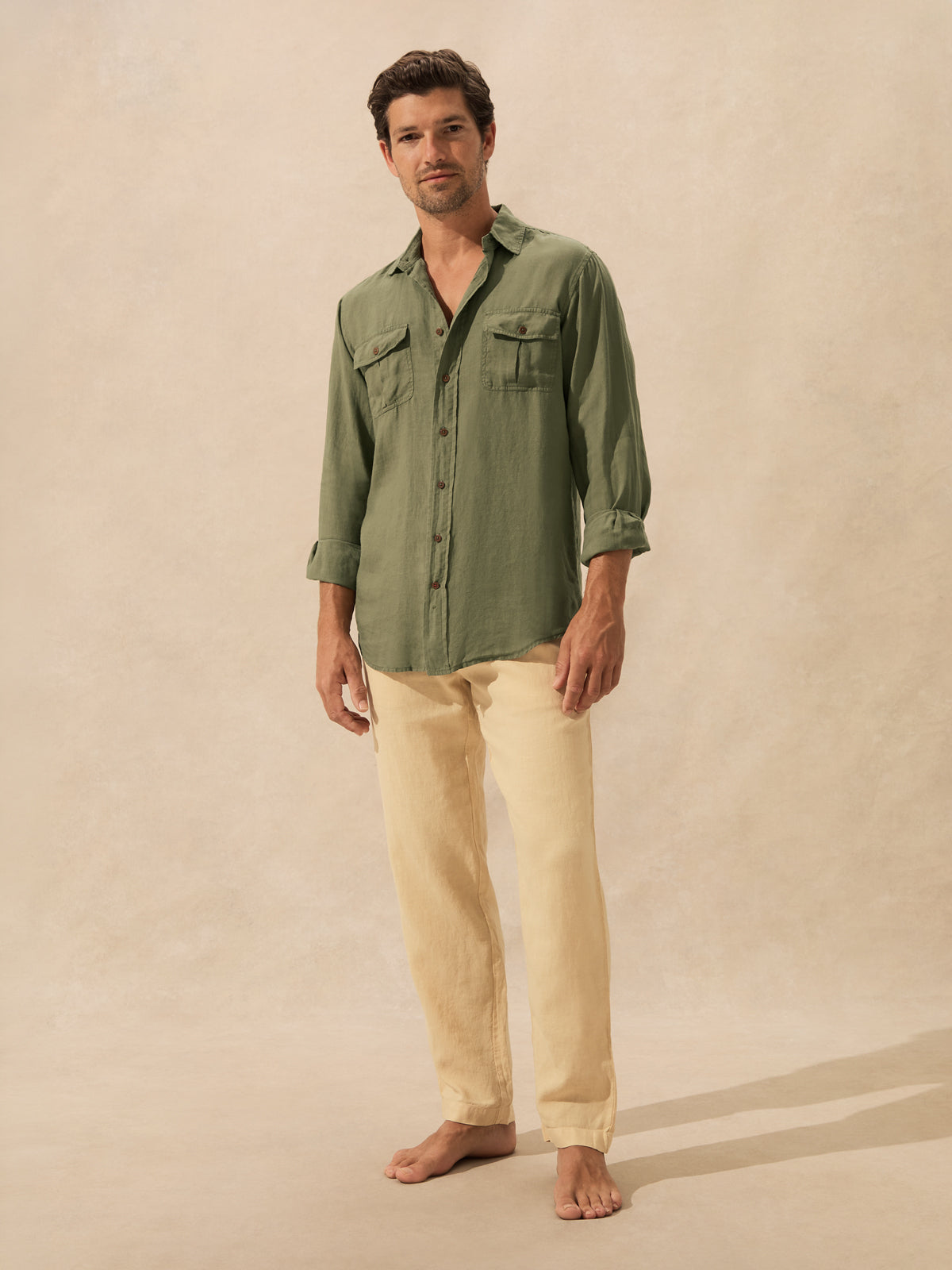 A man stands barefoot against a warm beige textured backdrop, wearing an olive green long-sleeve button-up shirt with chest flap pockets and light beige linen pants.