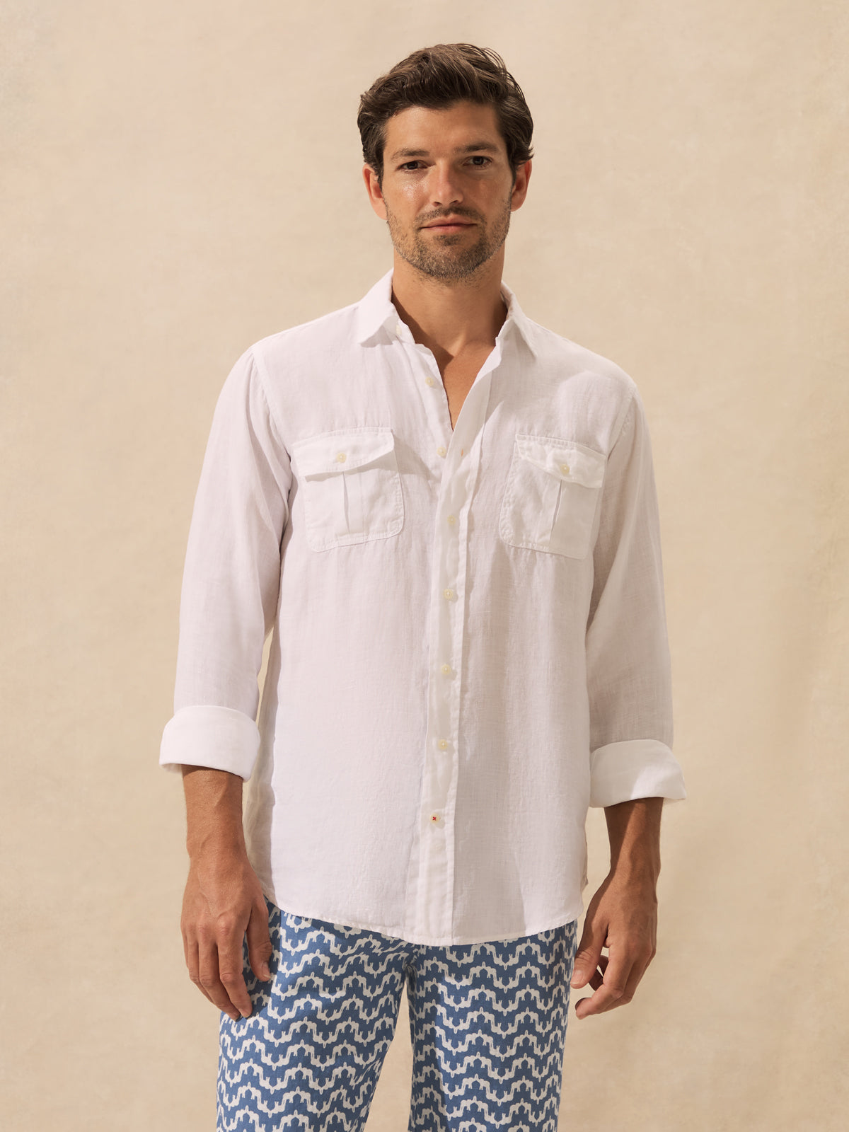 A man wearing a white long-sleeve button-down shirt with two chest pockets and blue patterned pants, standing against a plain beige studio background.