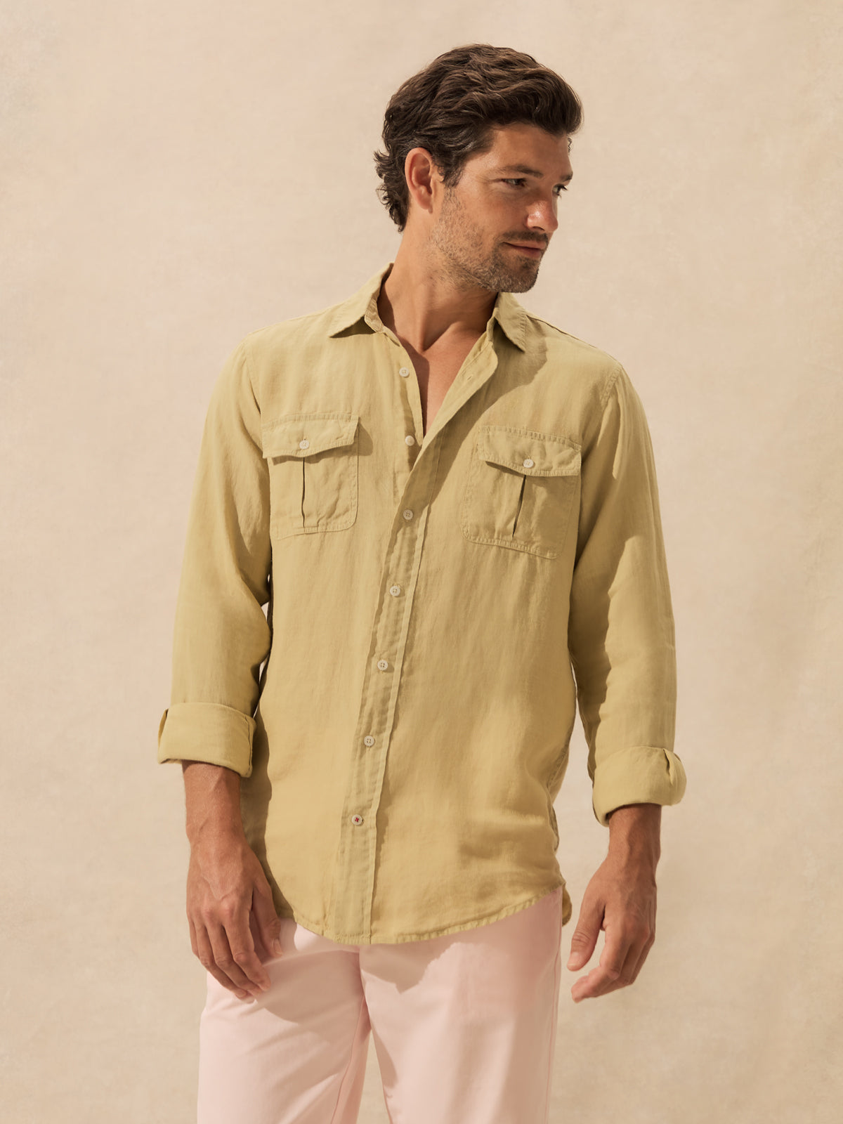 A man wearing a khaki long-sleeve button-up shirt with two chest pockets and light pink pants, standing against a textured beige background.