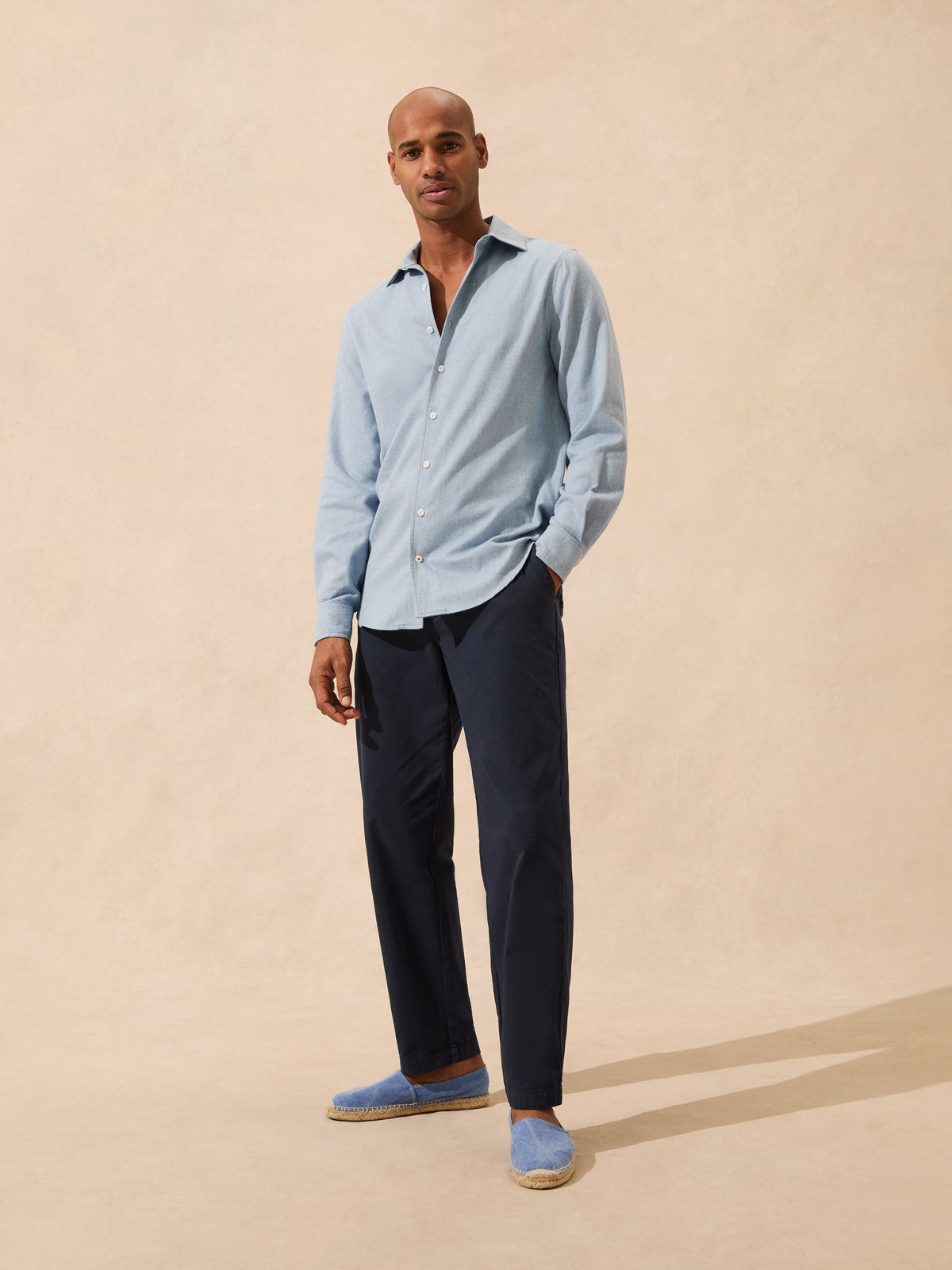 A man modeling a light blue long-sleeve button-up shirt, dark navy trousers, and light blue slip-on shoes against a warm beige textured studio backdrop.