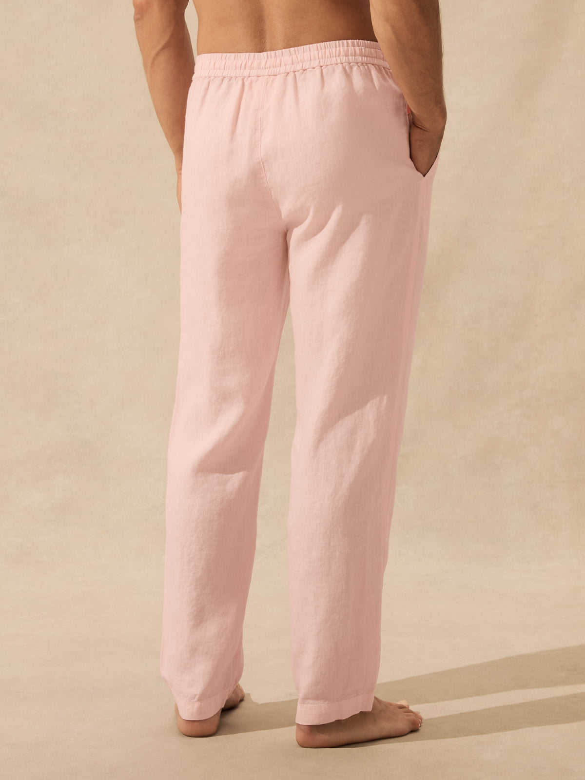 A person shown from the back wearing light pink lounge pants with an elastic waistband and side pockets, standing barefoot against a warm beige studio backdrop.