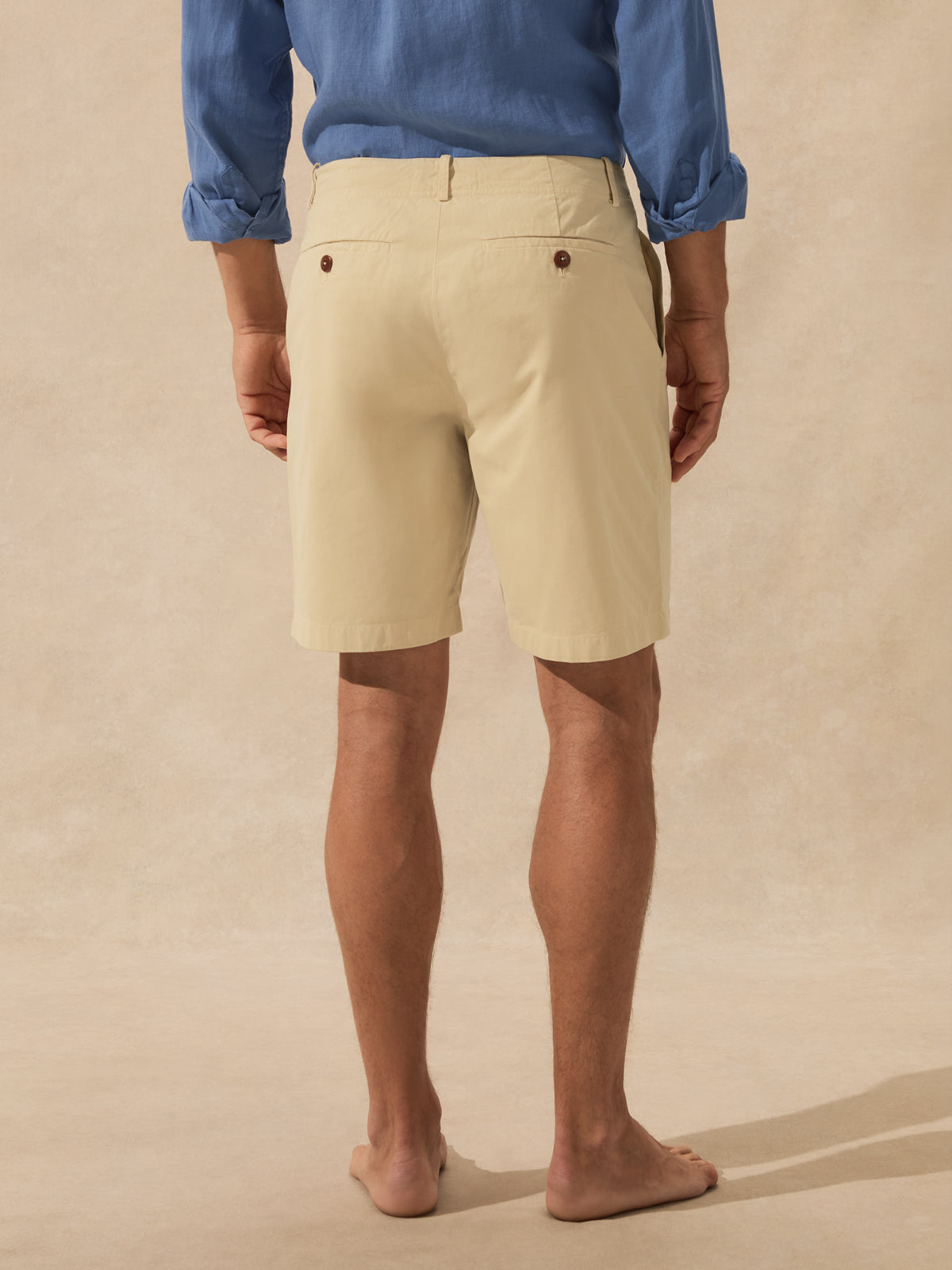 A man is shown from the back wearing light beige chino shorts standing barefoot against a textured beige studio backdrop.
