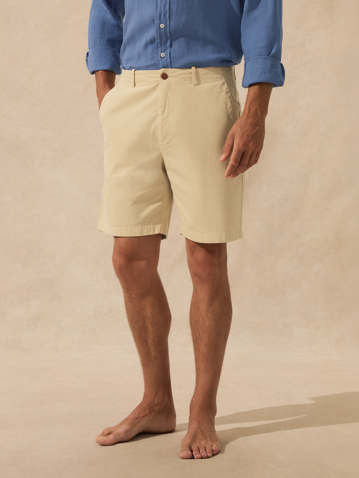 A man wearing beige chino shorts and a blue button-up shirt, standing barefoot against a textured beige studio backdrop.