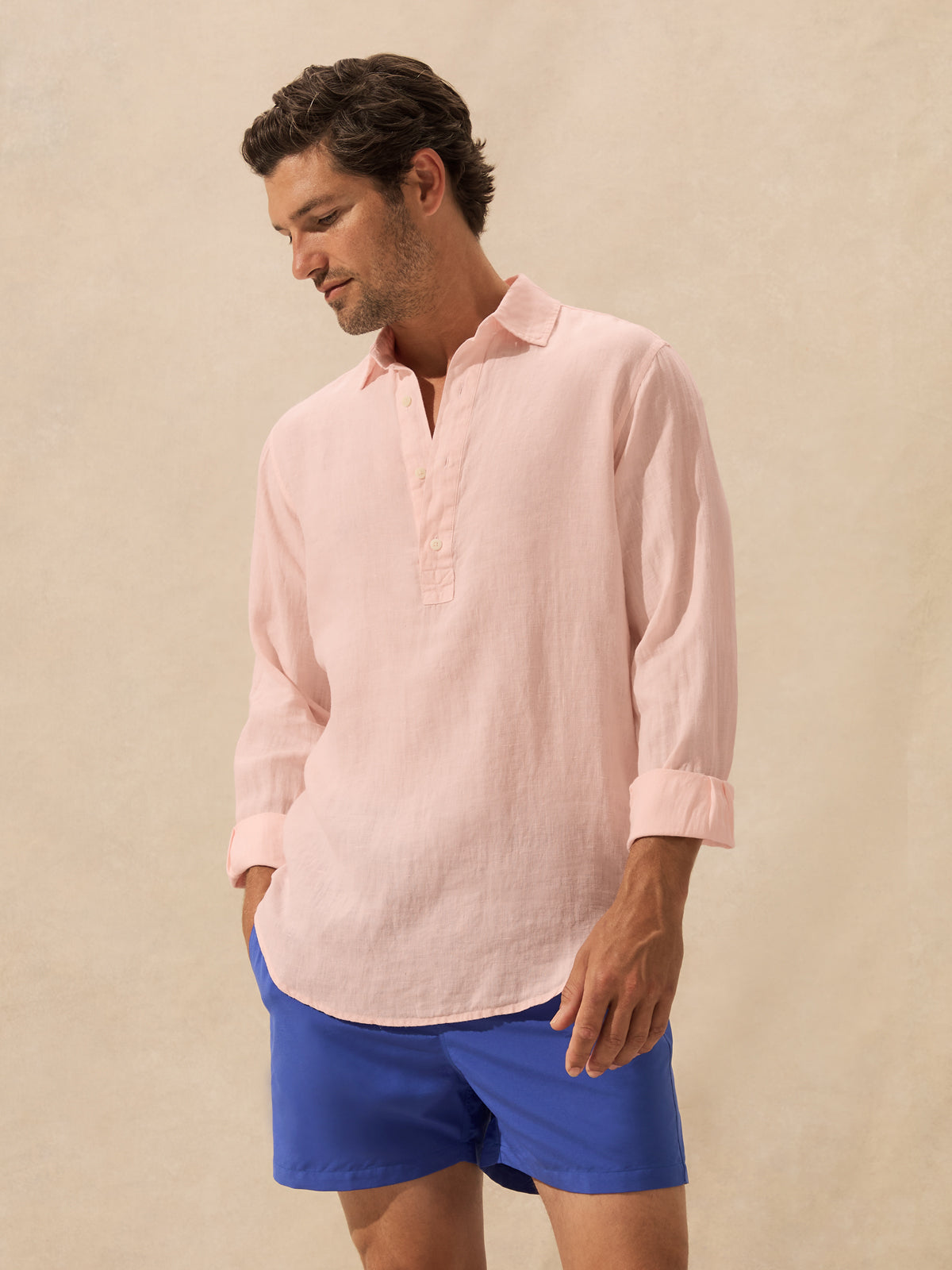 A man wearing a light pink long-sleeve linen henley shirt and royal blue shorts, standing against a textured beige studio backdrop.