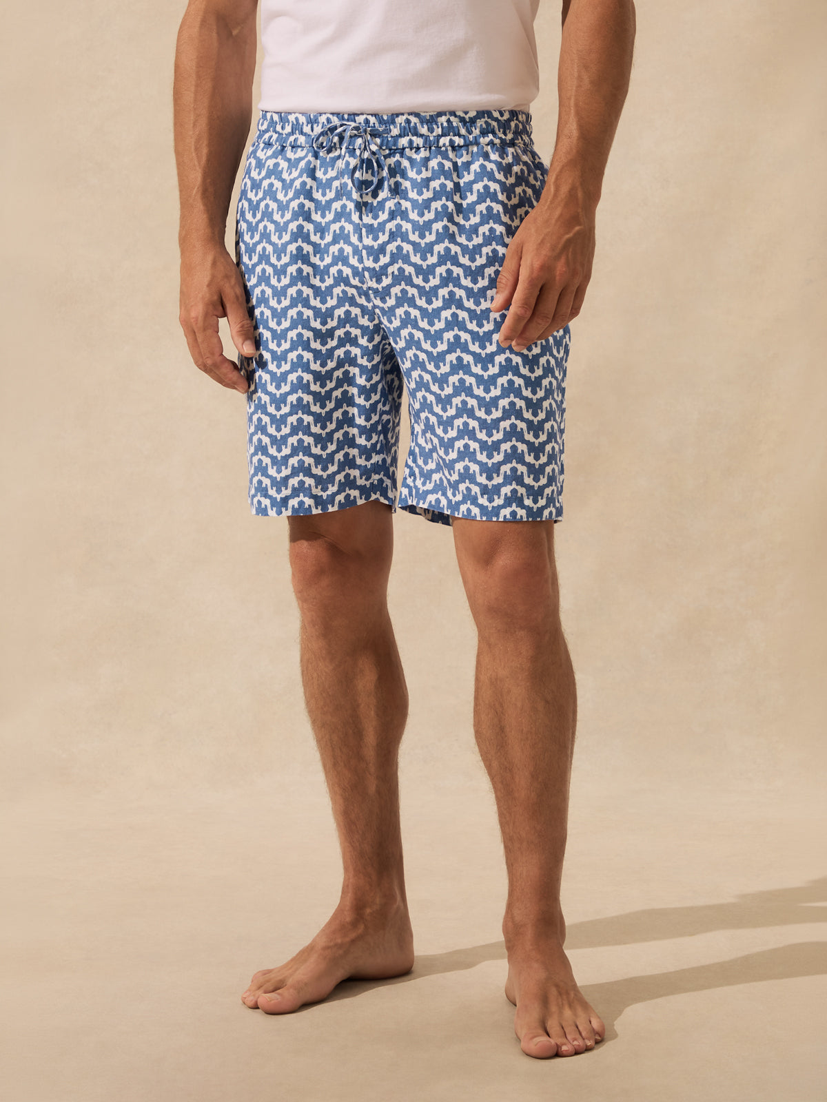 A man wearing blue and white patterned swim trunks and a white tank top, standing barefoot against a beige studio backdrop.