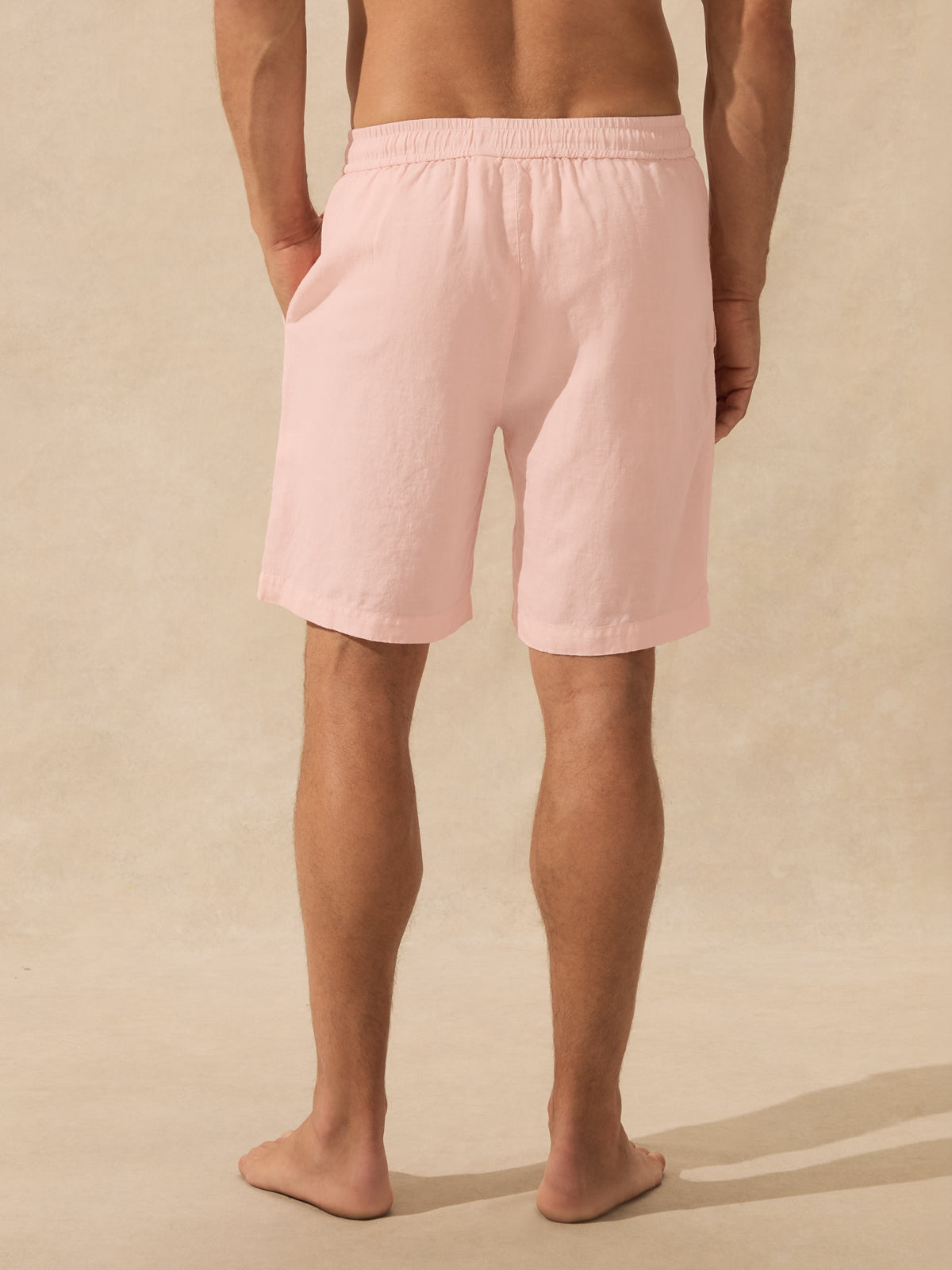 A man viewed from the back wearing light pink drawstring shorts, standing barefoot against a warm beige textured studio backdrop.