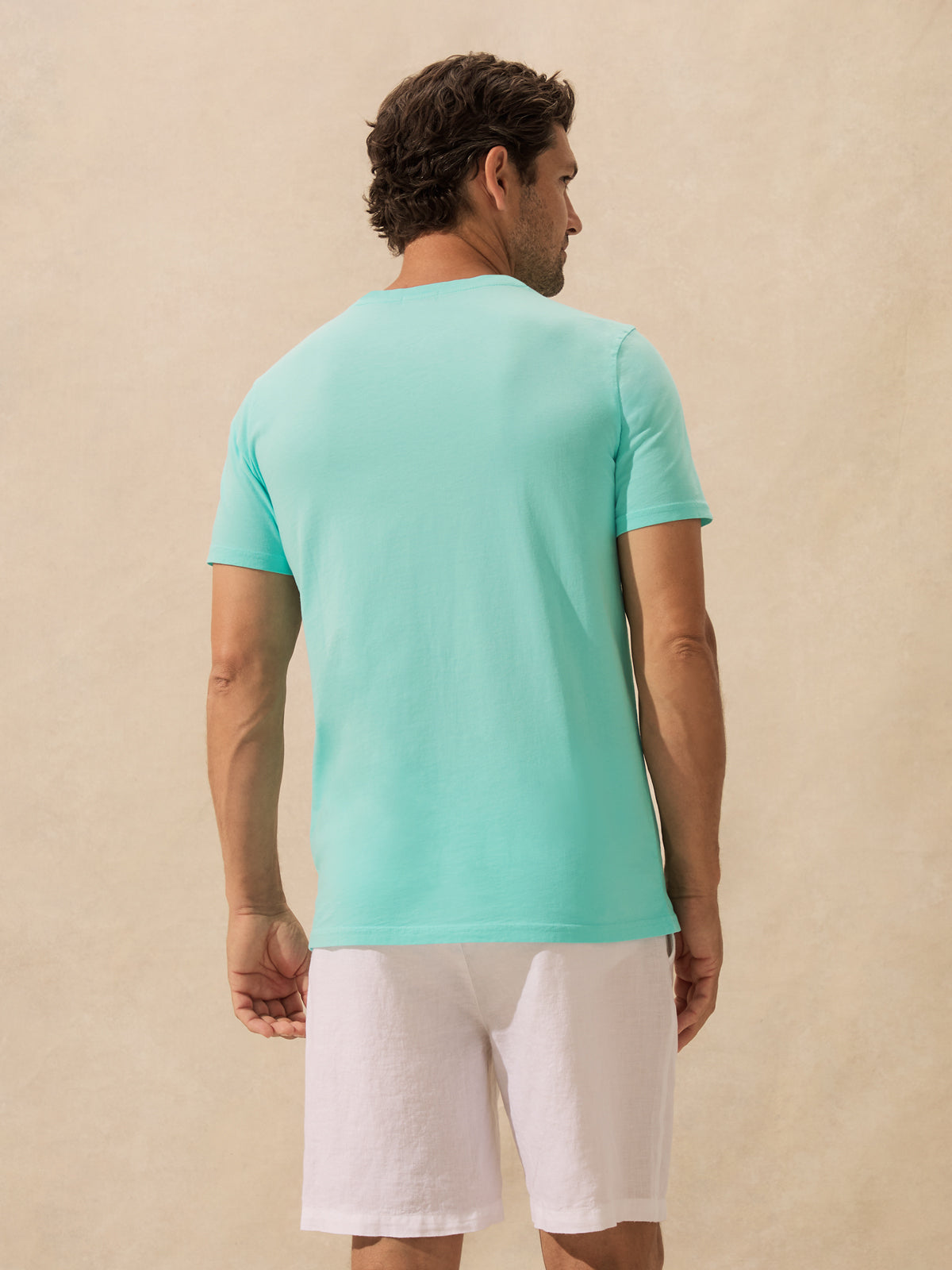 A man viewed from the back wearing a light mint green t-shirt and white linen shorts, standing against a textured beige wall.