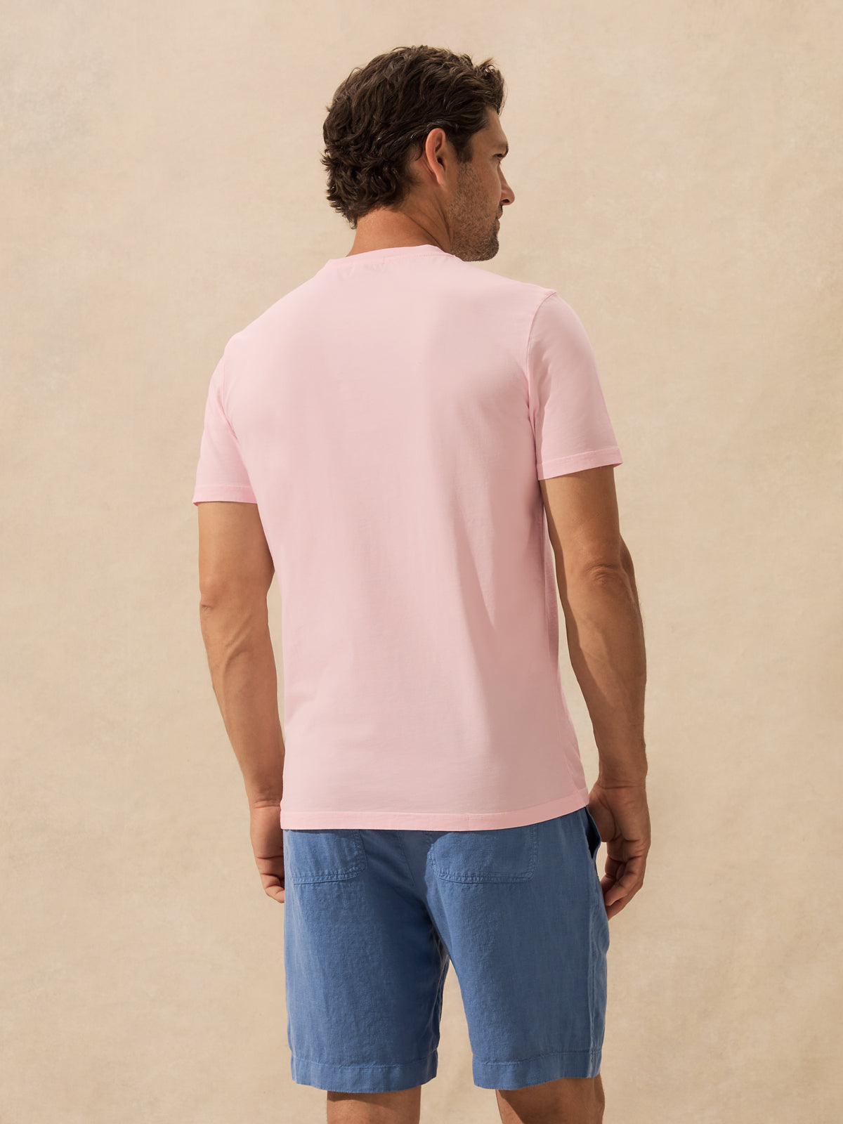 A man shown from the back wearing a light pink short-sleeve crewneck t-shirt and blue shorts, standing against a textured beige background.