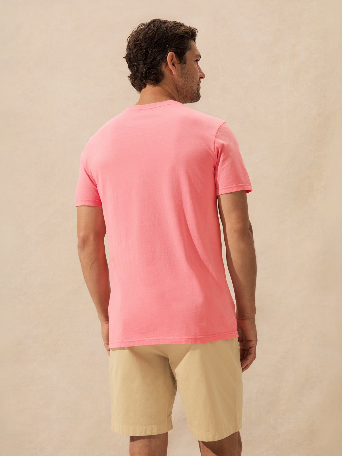A man shown from the back wearing a solid pink short-sleeve t-shirt and beige shorts, standing against a textured beige background.