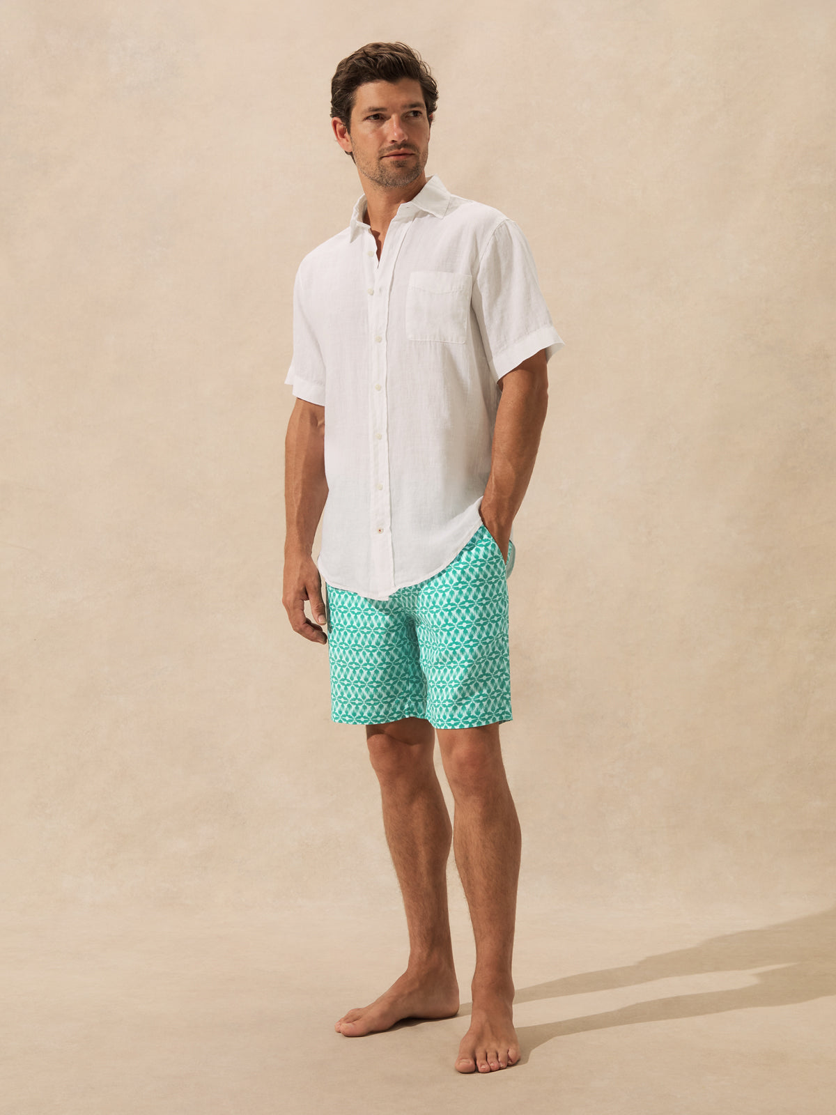 A man stands barefoot against a warm beige textured studio backdrop, wearing a white short-sleeve button-up shirt and turquoise shorts with a white geometric pattern.