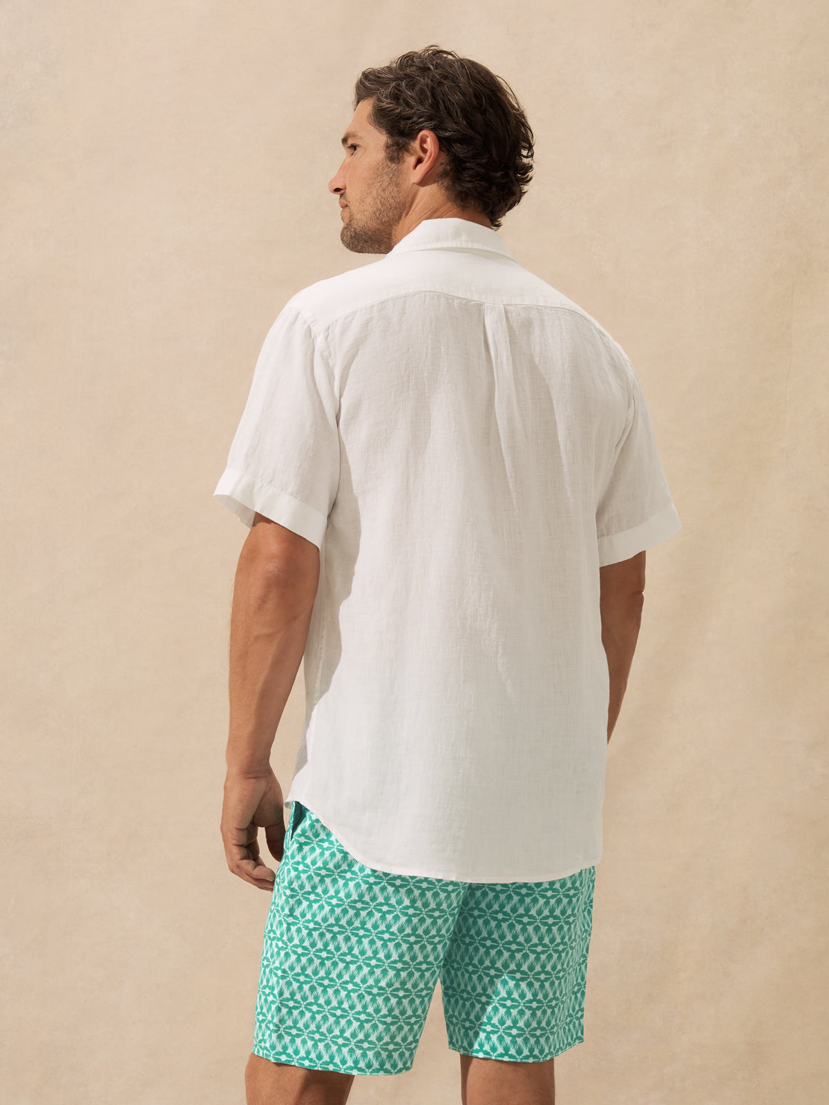 A man shown from the back wearing a white short-sleeve button-up shirt and teal patterned shorts against a warm beige textured background.