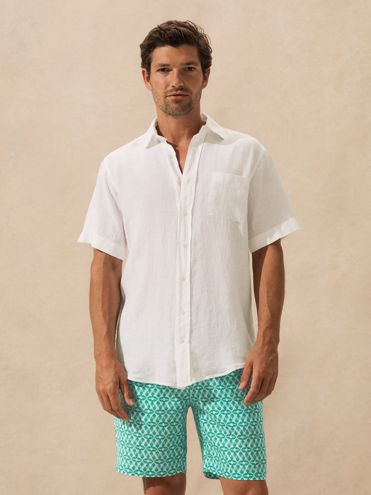A man wearing a white short-sleeve button-up shirt and teal patterned swim trunks, standing against a textured beige background.