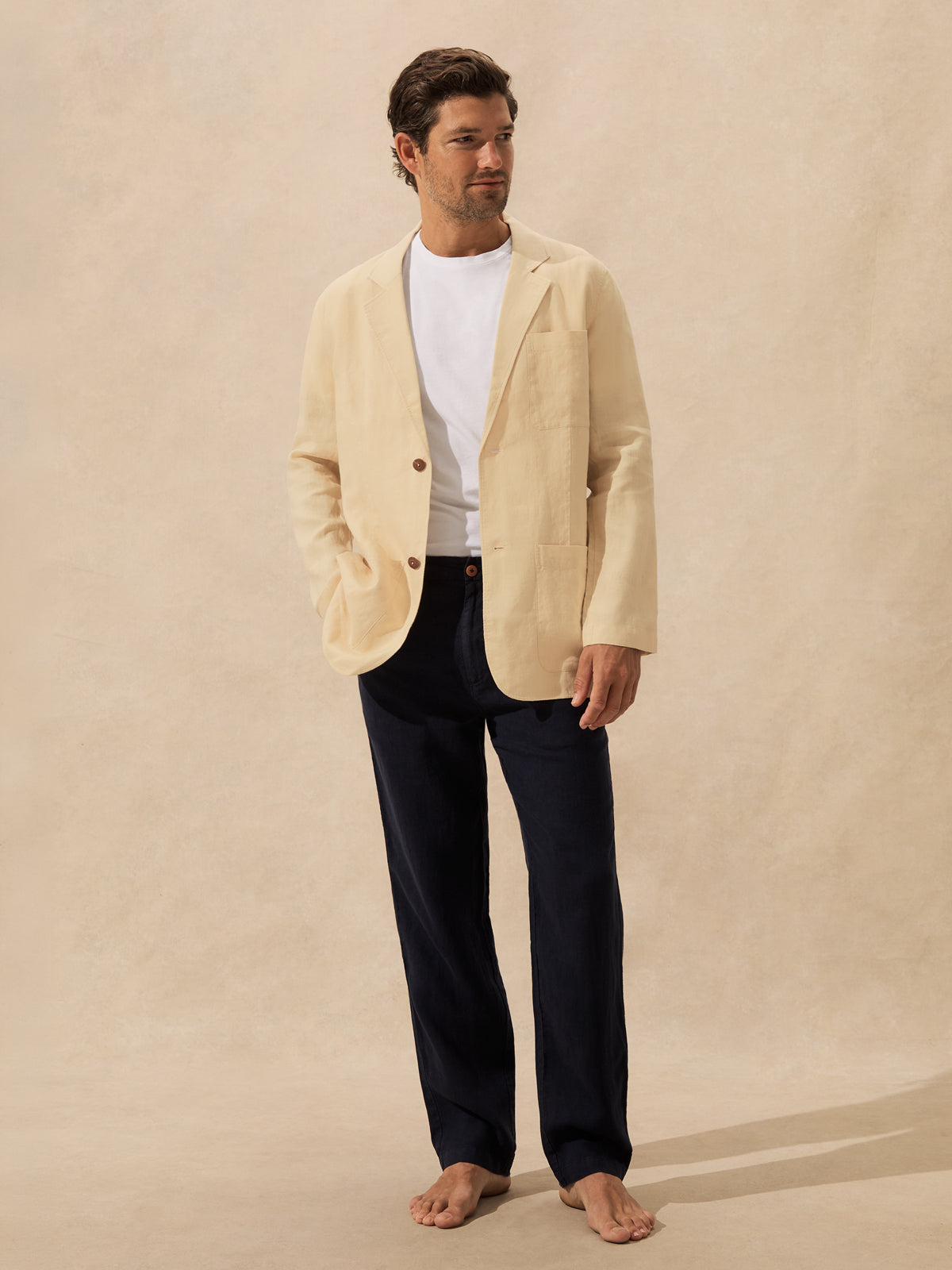 A man stands barefoot against a warm beige textured backdrop, wearing a light cream blazer over a white crew-neck t-shirt and dark navy trousers.