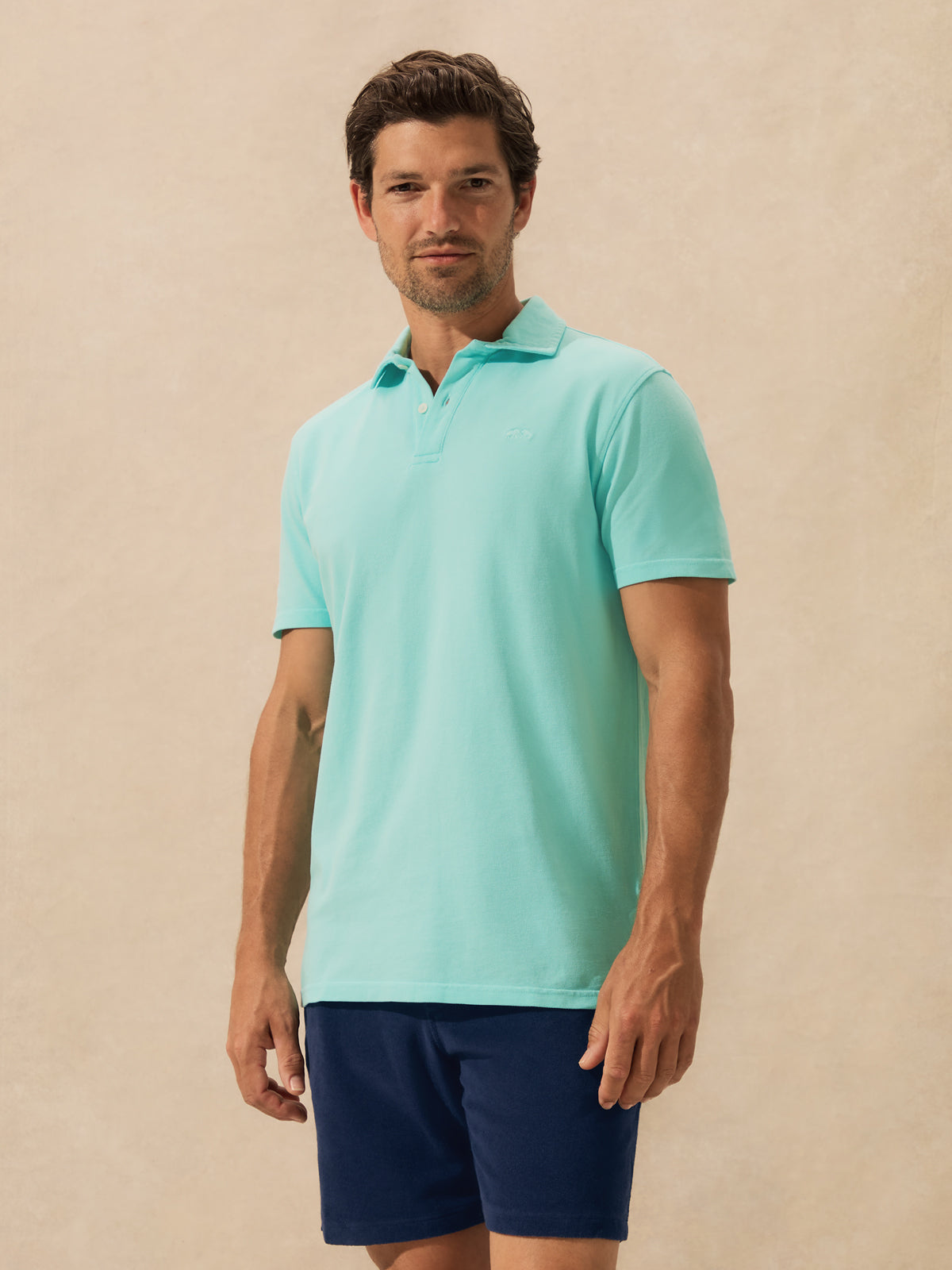 A man wearing a light mint green polo shirt and navy blue shorts, standing against a warm beige textured studio background.