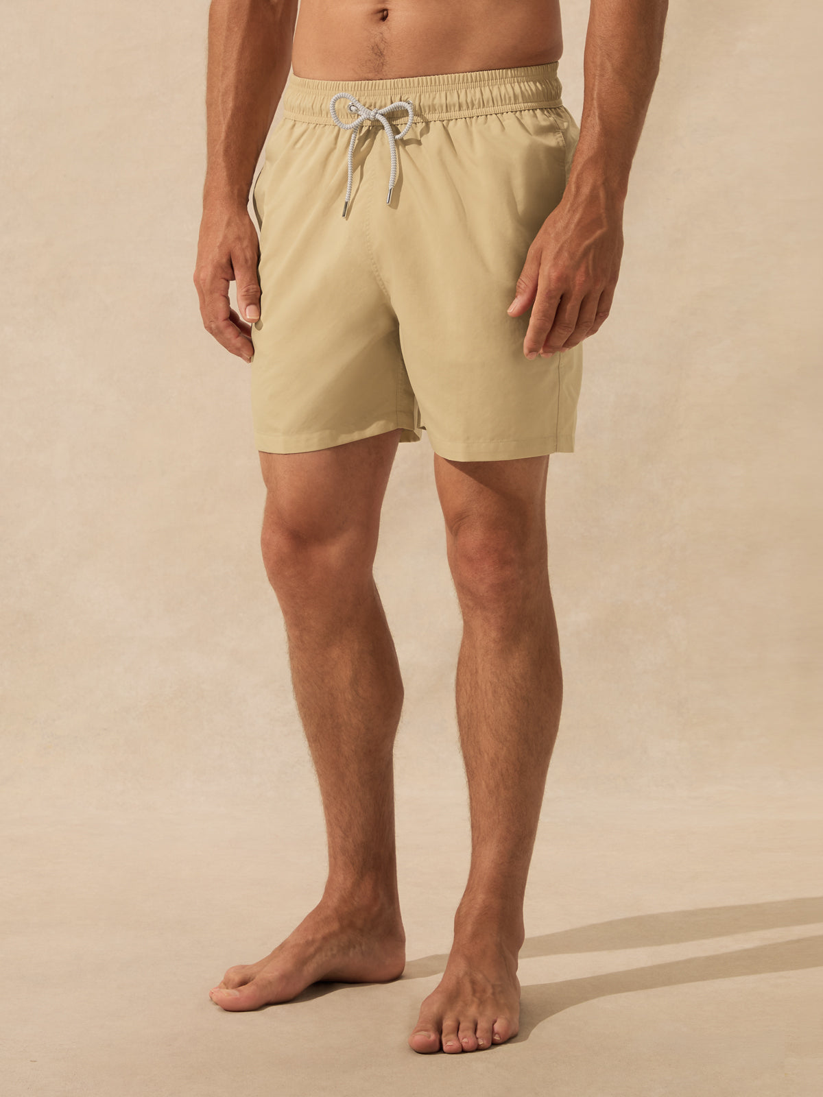 A man wearing beige swim trunks with a white drawstring, standing barefoot against a warm beige studio background.
