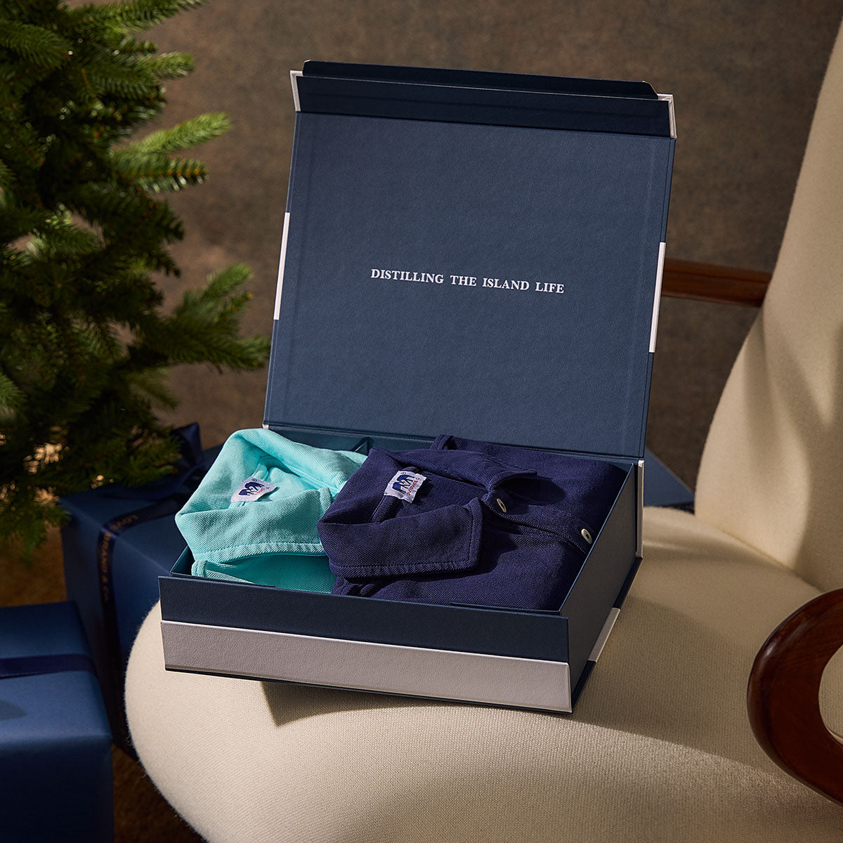 Open navy gift box with mens polo shirts on a beige surface, featuring 'Distilling the Island Life' text.