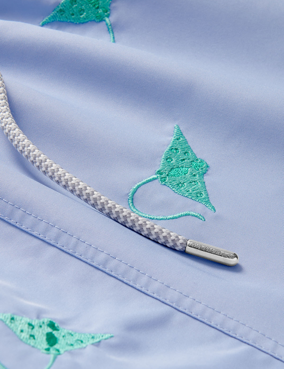 Close-up view of boys' swim shorts featuring vibrant turquoise manta ray embroidery on a pastel blue quick-dry fabric, with a detailed braided drawstring.