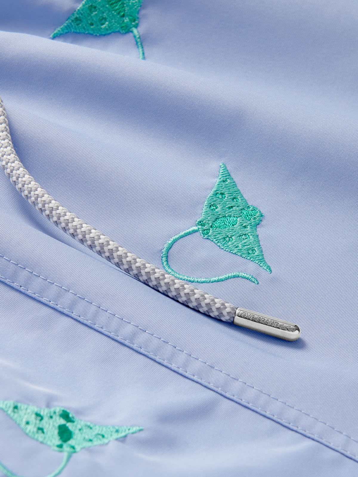 Close-up view of boys' swim shorts featuring vibrant turquoise manta ray embroidery on a pastel blue quick-dry fabric, with a detailed braided drawstring.