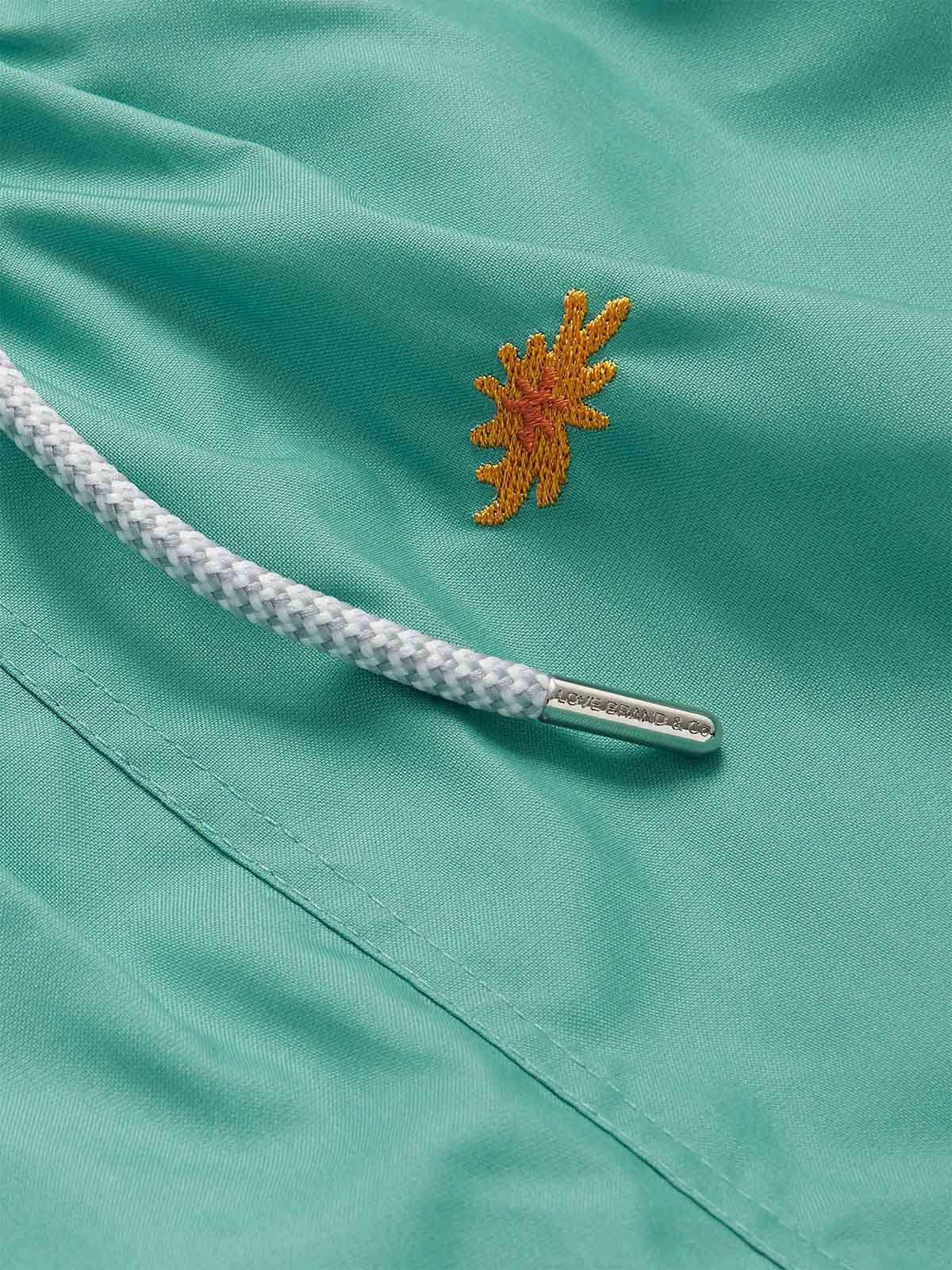 Close-up of the Men's Primavera Embroidered Staniel Swim Shorts in Riviera green. Embroidered swim shorts featuring an embroidered orange sea flower and drawstring detail.