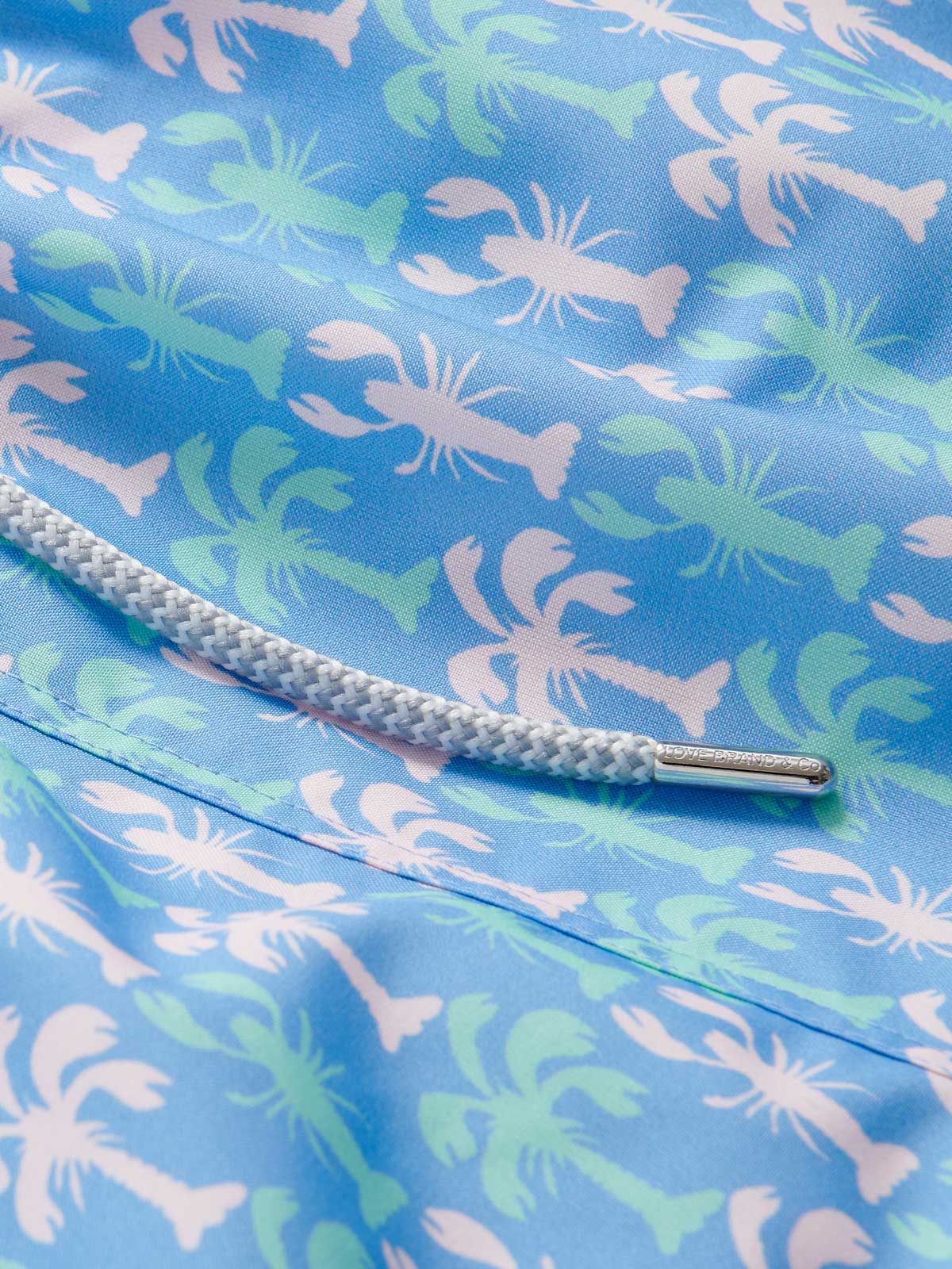 Close-up of Men's Party Like a LobStar Staniel Swim Shorts featuring a pattern of dancing lobsters and swaying palm trees on a blue background with a white drawstring.