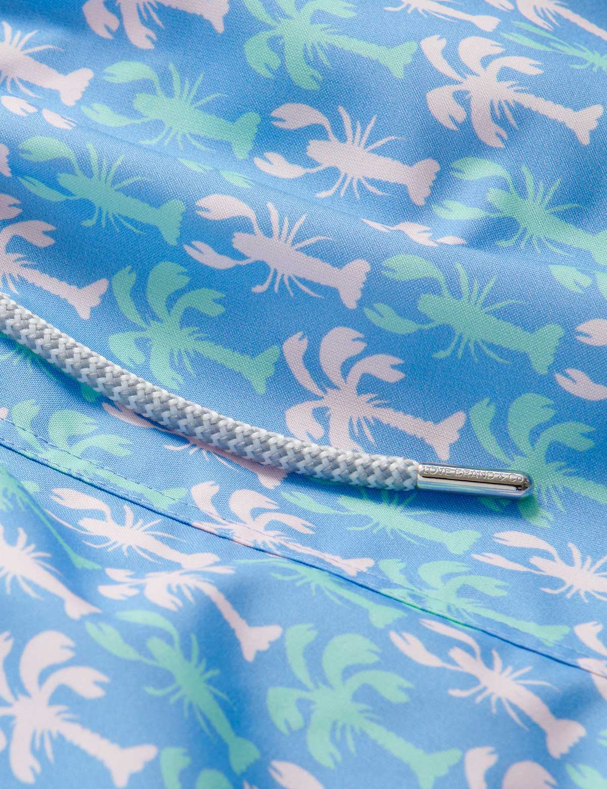 Close-up of Men's Party Like a LobStar Staniel Swim Shorts featuring a pattern of dancing lobsters and swaying palm trees on a blue background with a white drawstring.