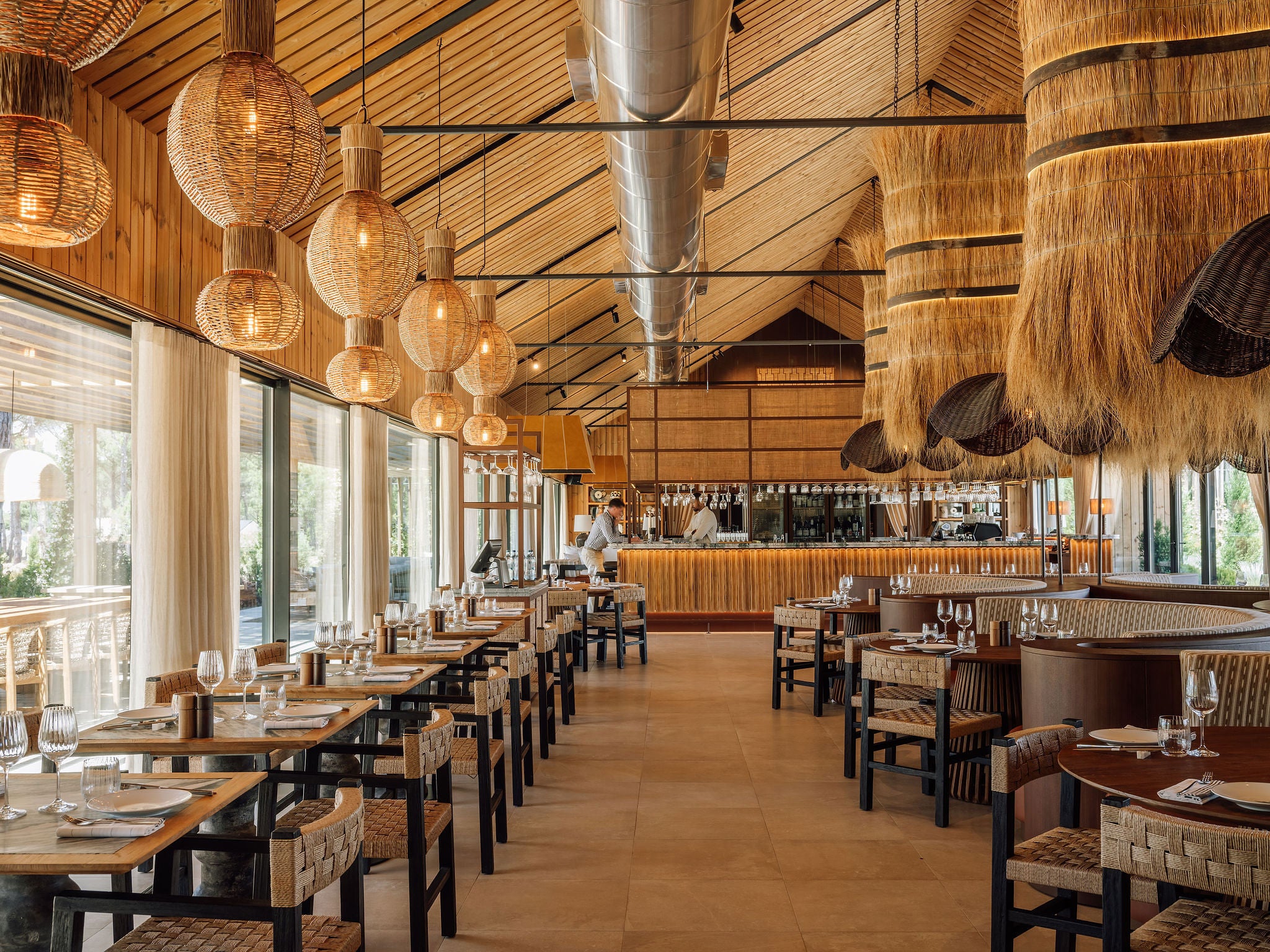 A long, high-ceilinged restaurant interior with warm wood walls, large woven pendant lights, thatched hanging decor, and rows of dining tables and chairs.