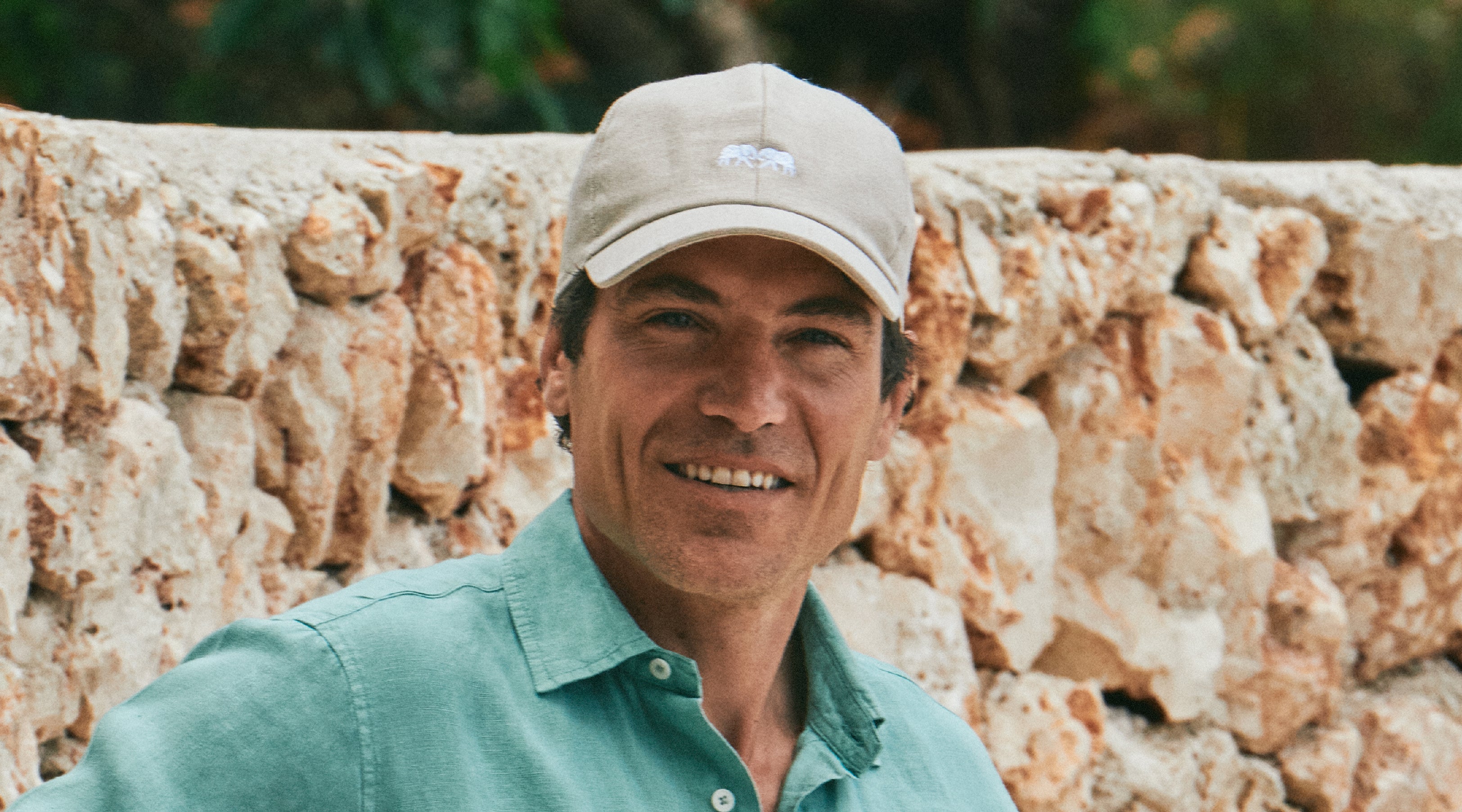 Man wearing a beige baseball cap and green beach shirt standing in front of a stone wall.