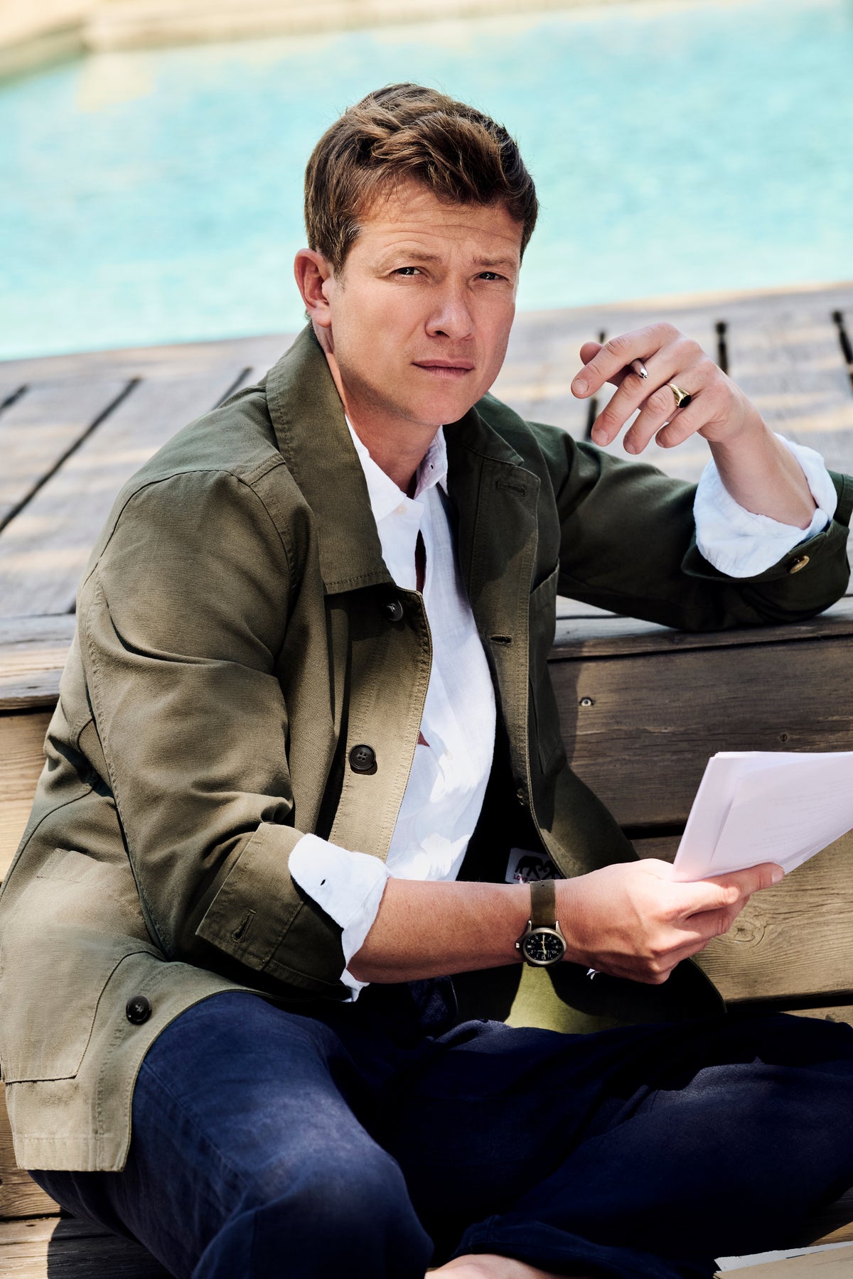 Ed Speleers sitting outdoors by a pool wearing an olive green jacket over a white shirt and dark blue trousers, holding a piece of paper.