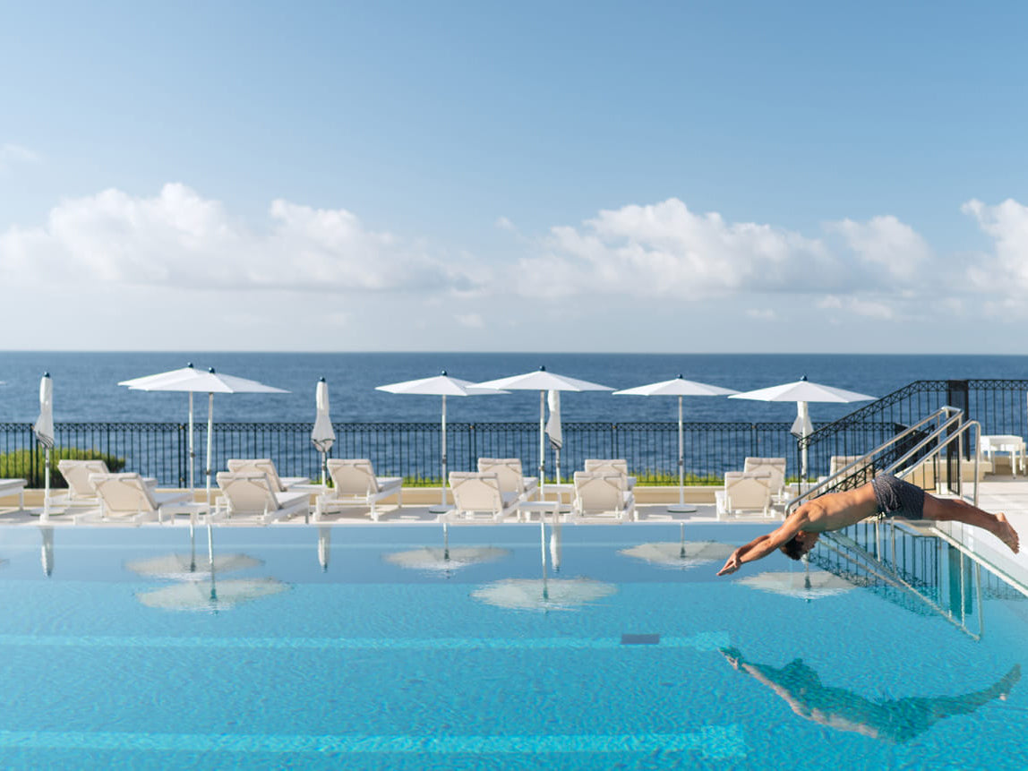 Person diving into a pool with ocean view and sun umbrellas