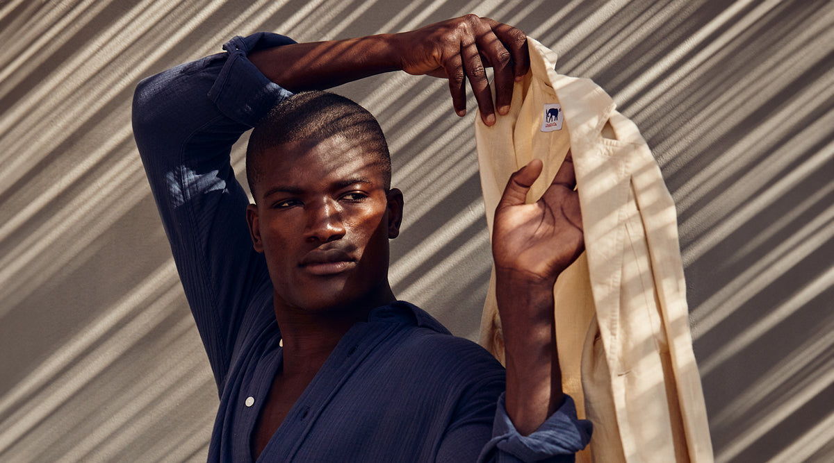 A male model wearing a dark navy blue shirt holds up a light beige shirt against a background with diagonal shadows.