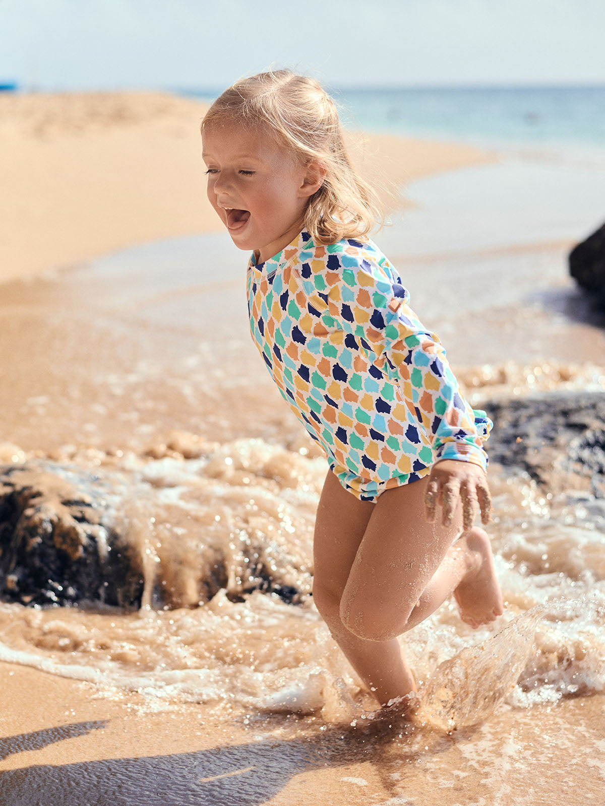 Child in a colourful rash vest and bikini bottoms playing in the water at the beach