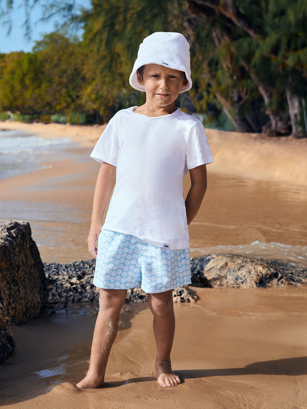 Child standing on a beach wearing a white t-shirt, light blue swim shorts, and a white bucket hat.