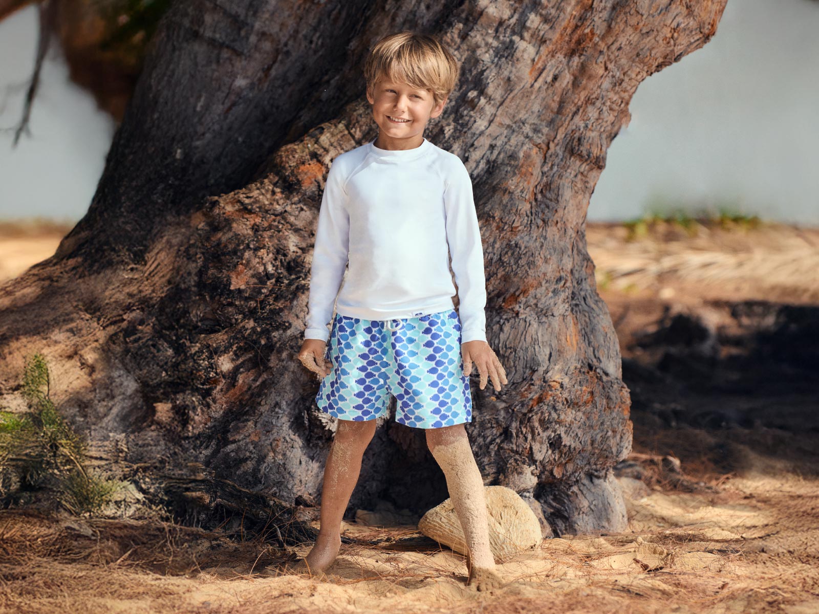 Child wearing a white rash vest and blue patterned swim shorts standing in front of a large tree.