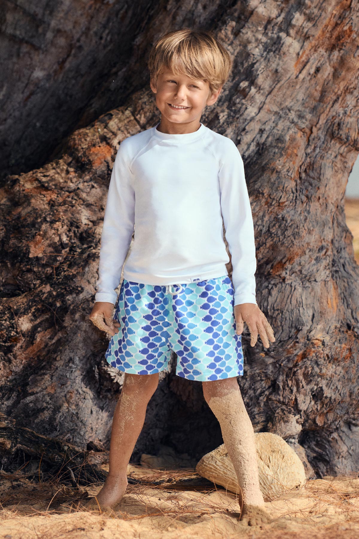 Child wearing a white long-sleeve rash vest and blue patterned swim shorts standing in front of a large tree.