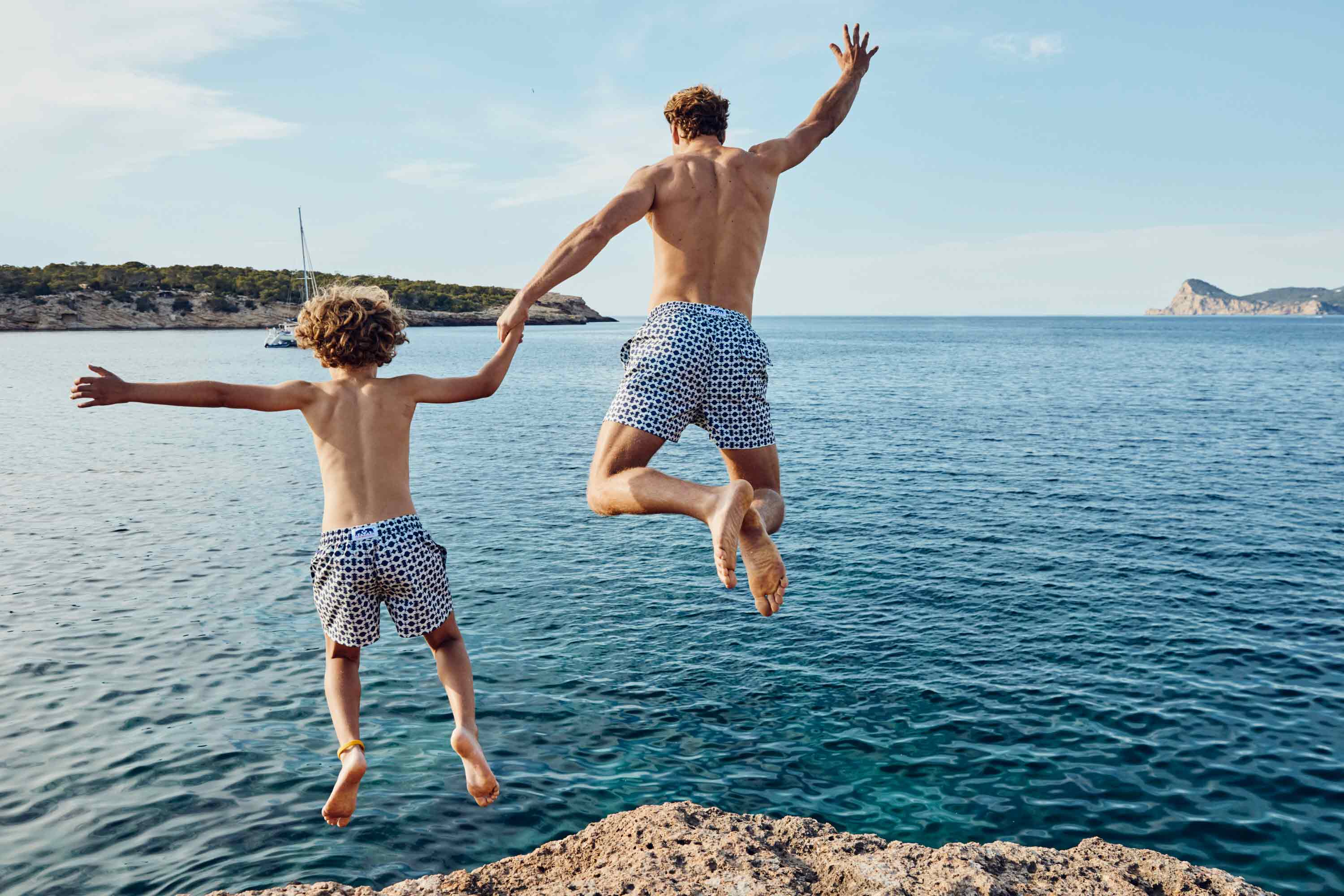 Two people jumping into the ocean from a cliff with a clear blue sky.