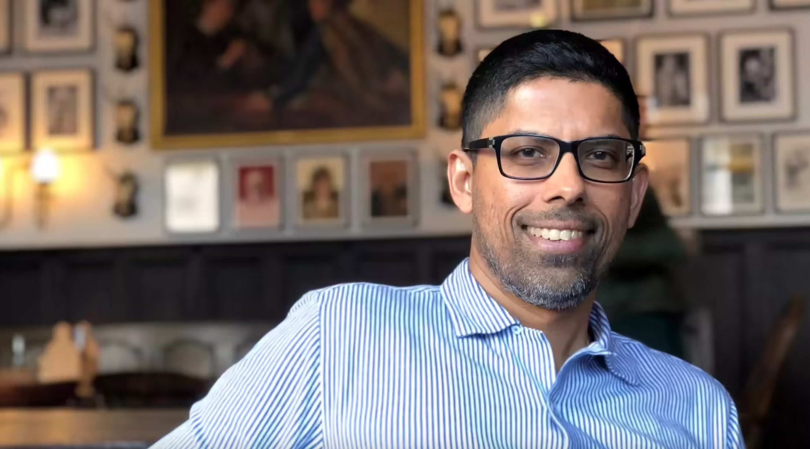 Man wearing glasses and a blue striped shirt in a room with framed pictures on the wall.