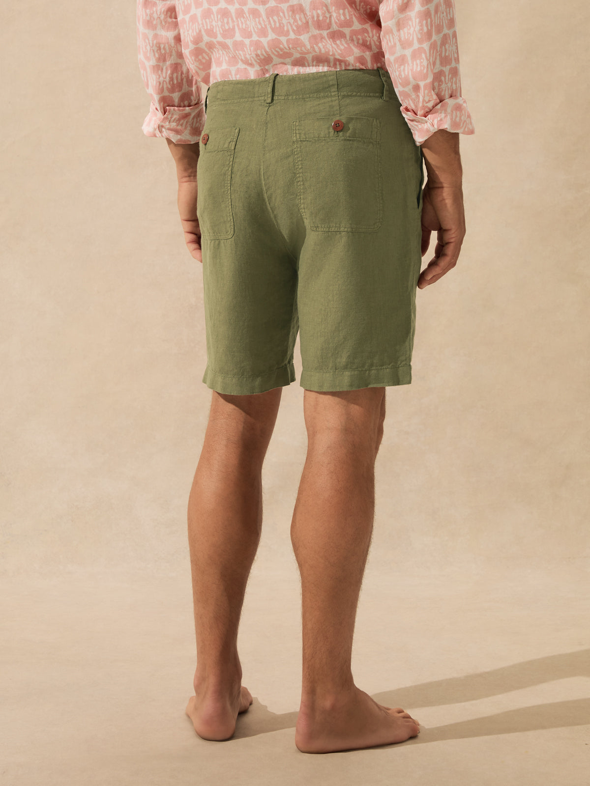 A person shown from the back wearing olive green linen shorts, standing barefoot against a textured beige studio backdrop.