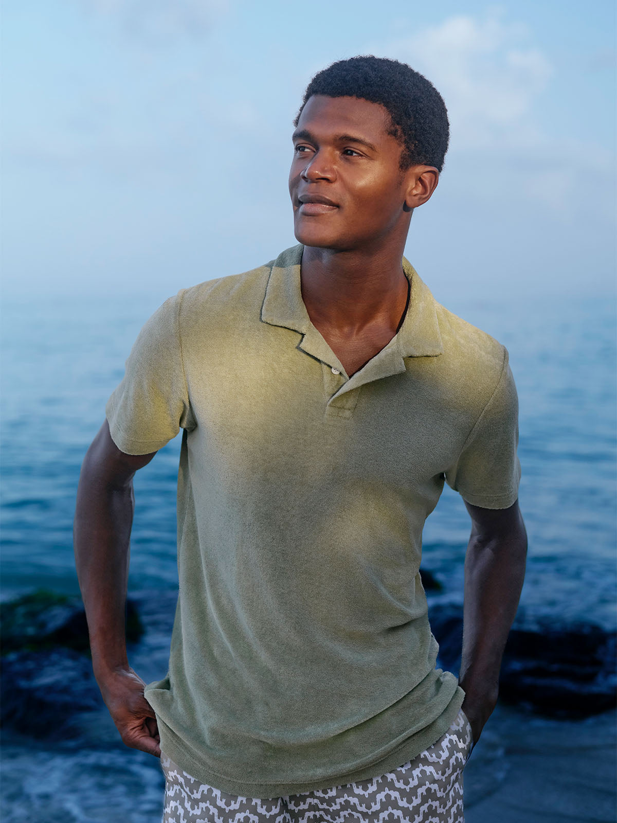 Man wearing a light green polo shirt and patterned swim shorts standing by the ocean.