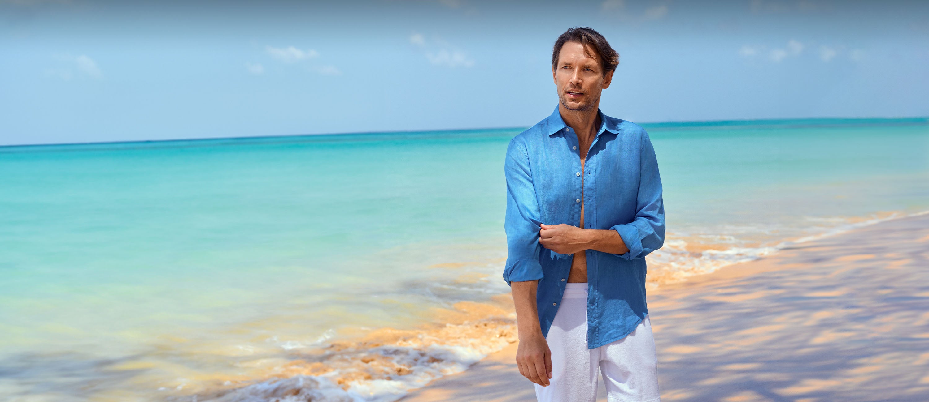 Man standing on a beach wearing mens designer beachwear with clear blue water and sky
