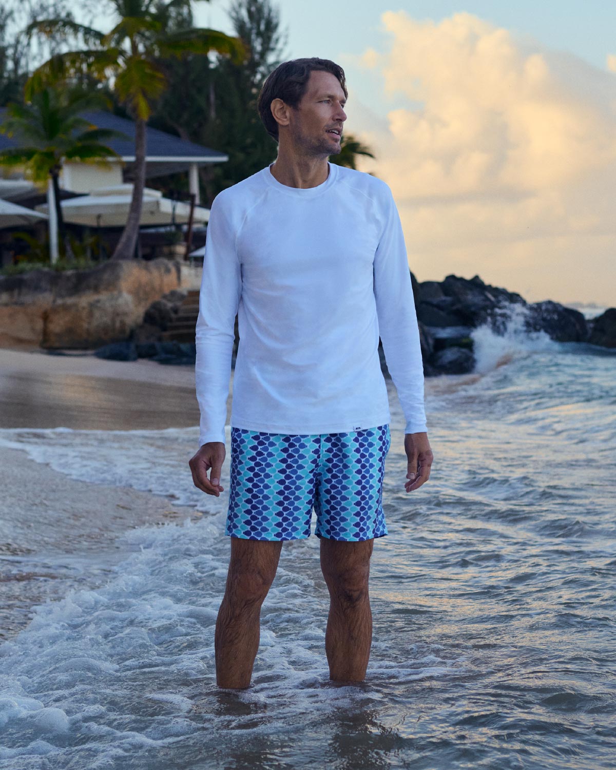 Man standing in shallow water at the beach wearing a white long-sleeve rash vest and blue patterned swim shorts.