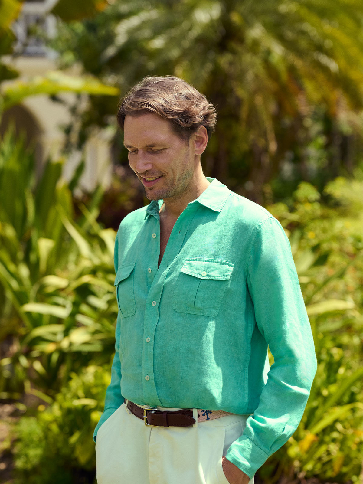 Man wearing a green shirt and white pants standing in a tropical setting with greenery.