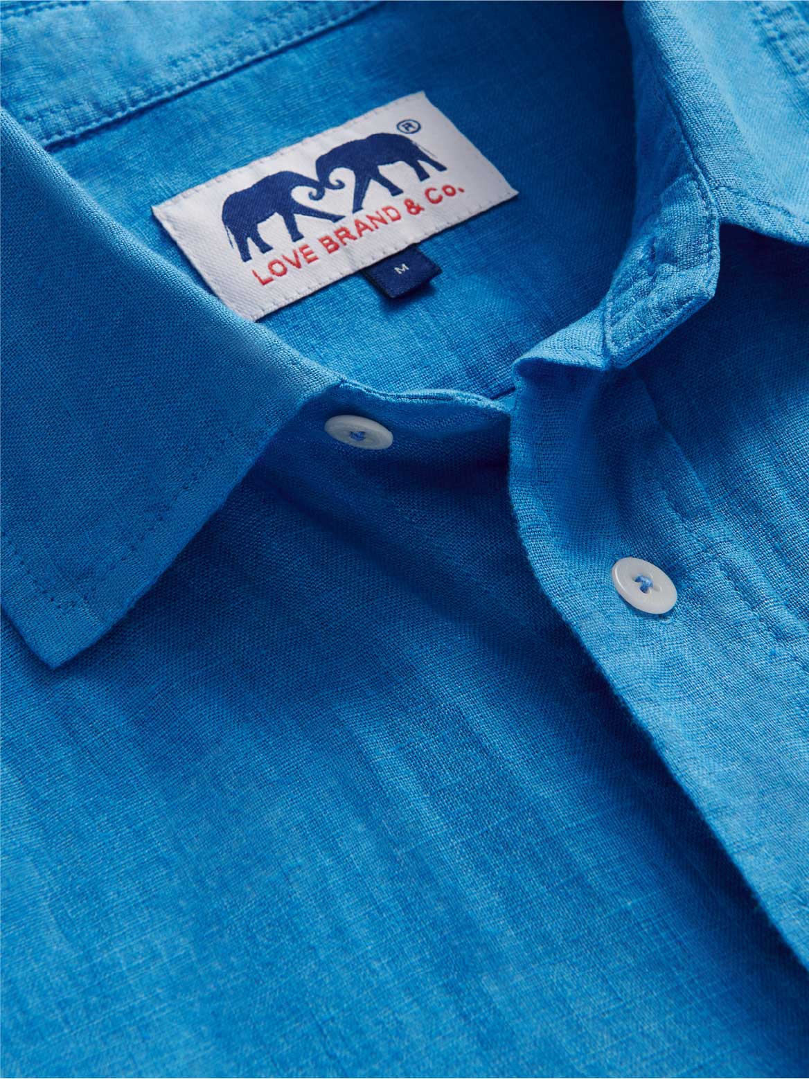 Close-up of a blue linen shirt with a love brand label featuring two elephants and text.