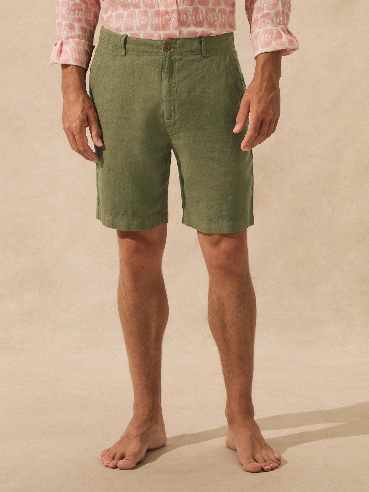 A man wearing olive green linen shorts, a pink patterned shirt, and standing barefoot against a beige studio backdrop.